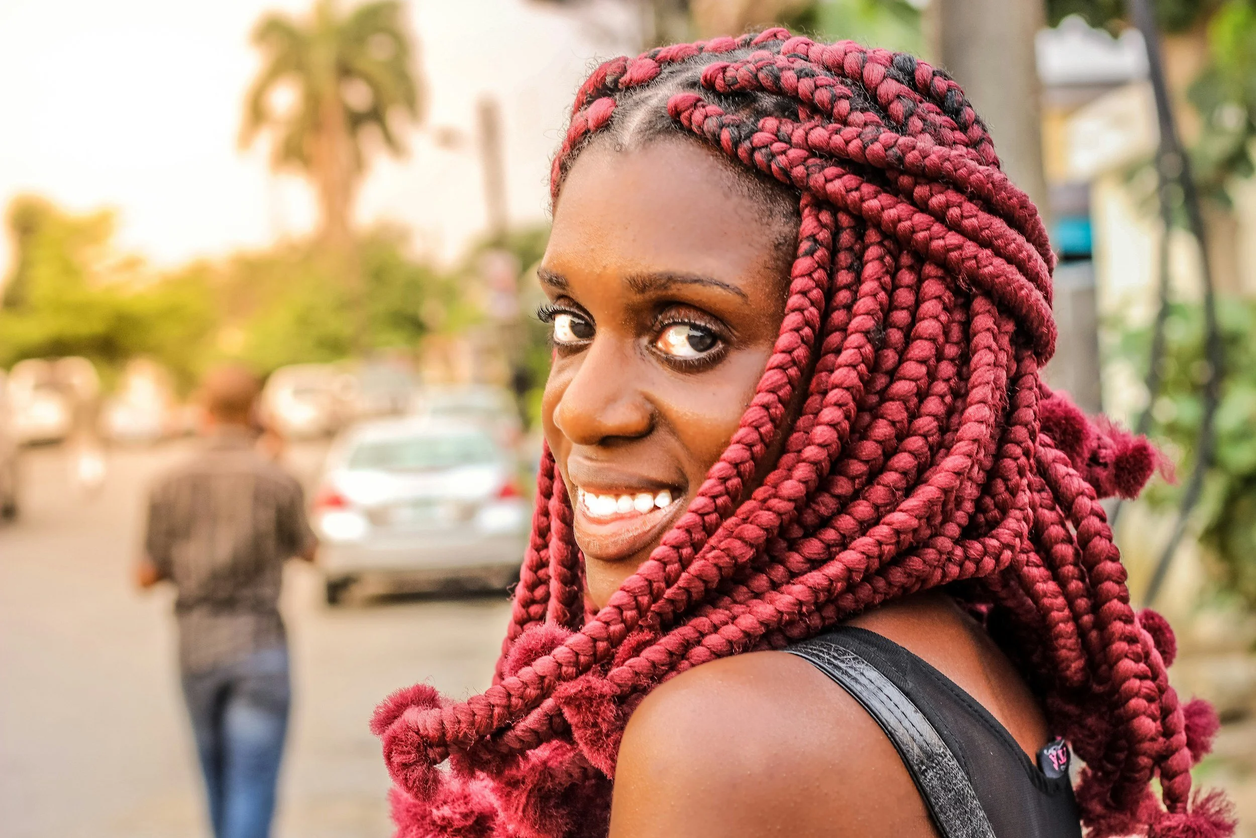 Close-up of a woman with dark skin and red braided hair smiling outdoors.