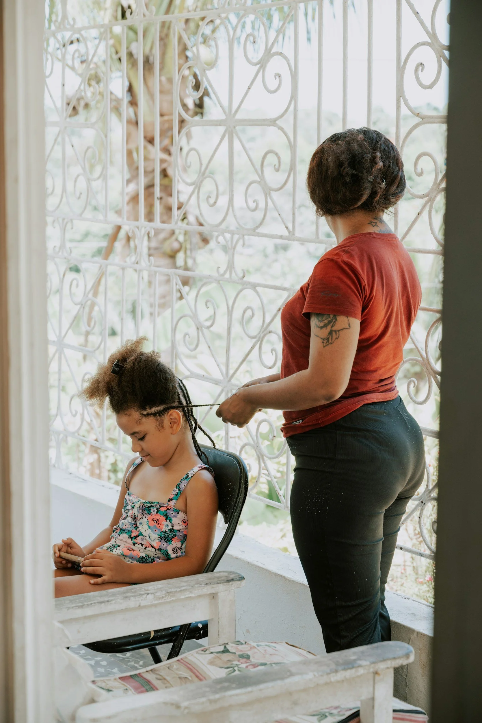 A woman with short wavy hair and tattoos on her arm is standing behind a young girl with braided hair, sitting on a chair, on a porch with a decorative white metal railing, while the girl looks at her phone.