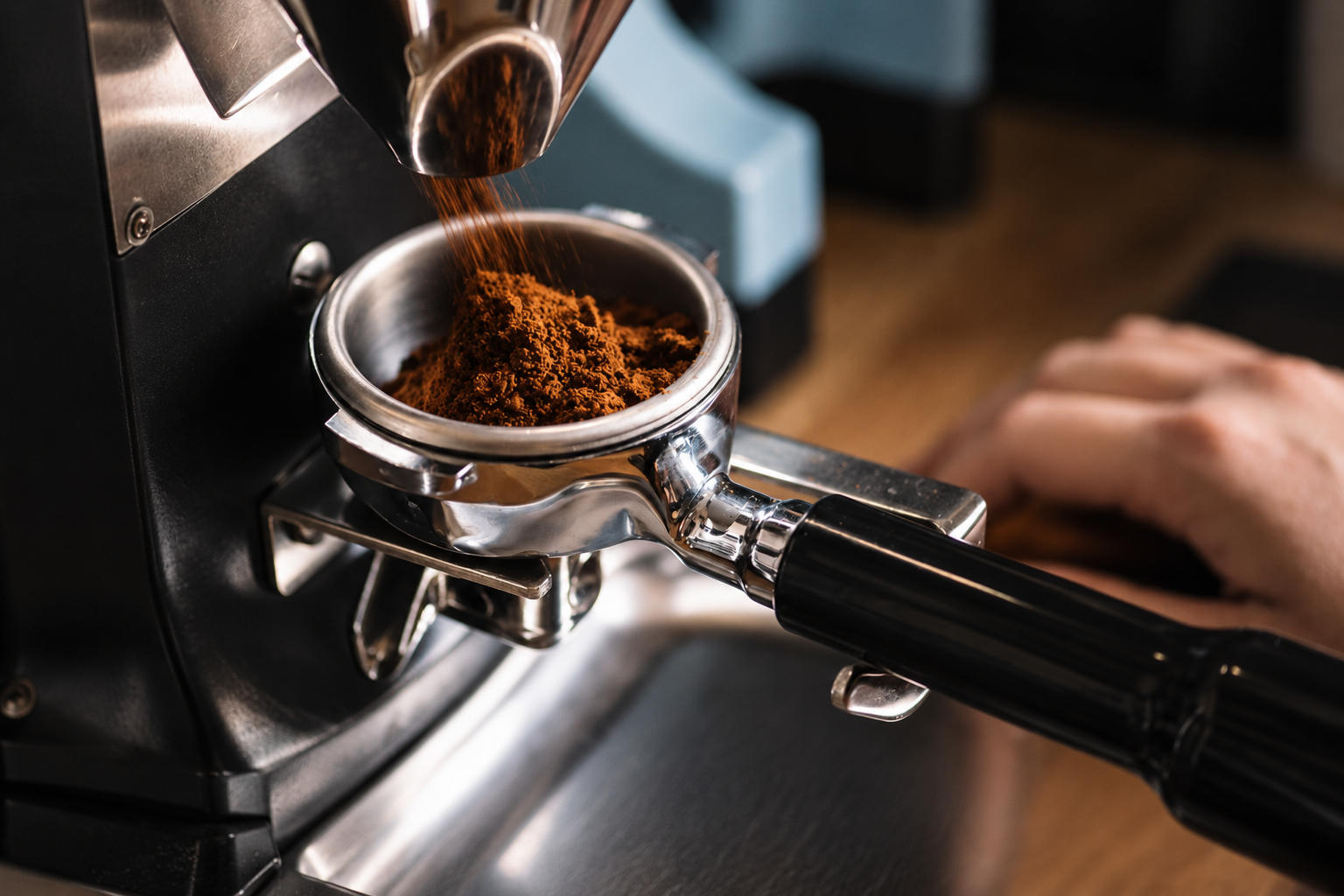 Close-up of a portafilter filled with ground coffee, attached to an espresso machine, with coffee grounds falling into it from a scoop.
