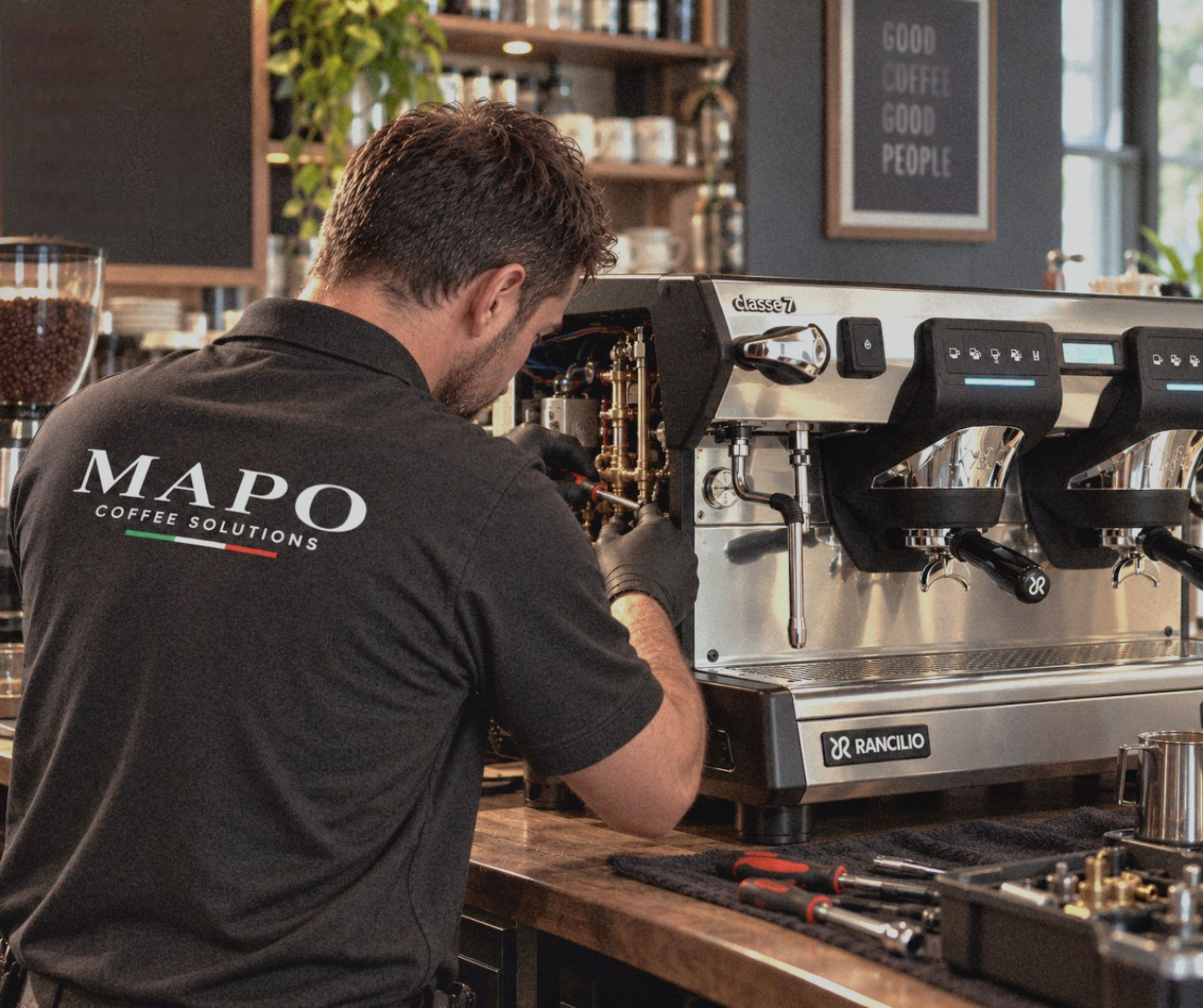 A man working on a commercial coffee machine in a coffee shop with a chalkboard sign reading 'Good Coffee Good People' in the background.