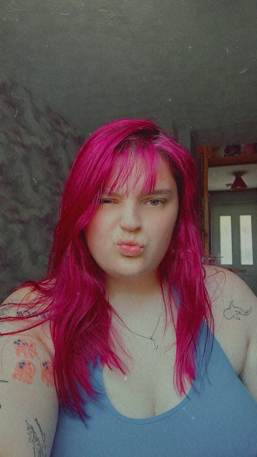 Person with long, neon pink hair, wearing a blue tank top, taking a selfie indoors with tattoos on arms and a nose ring, making a pouty face.