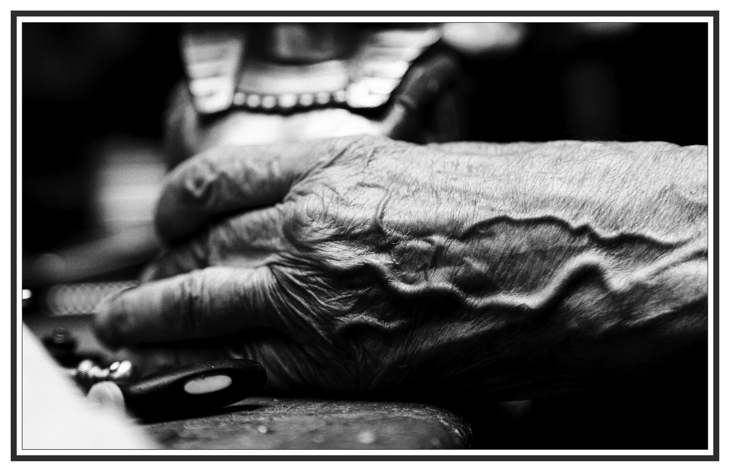 Close-up of an elderly person's hand resting on a surface, with a blurred person in the background.