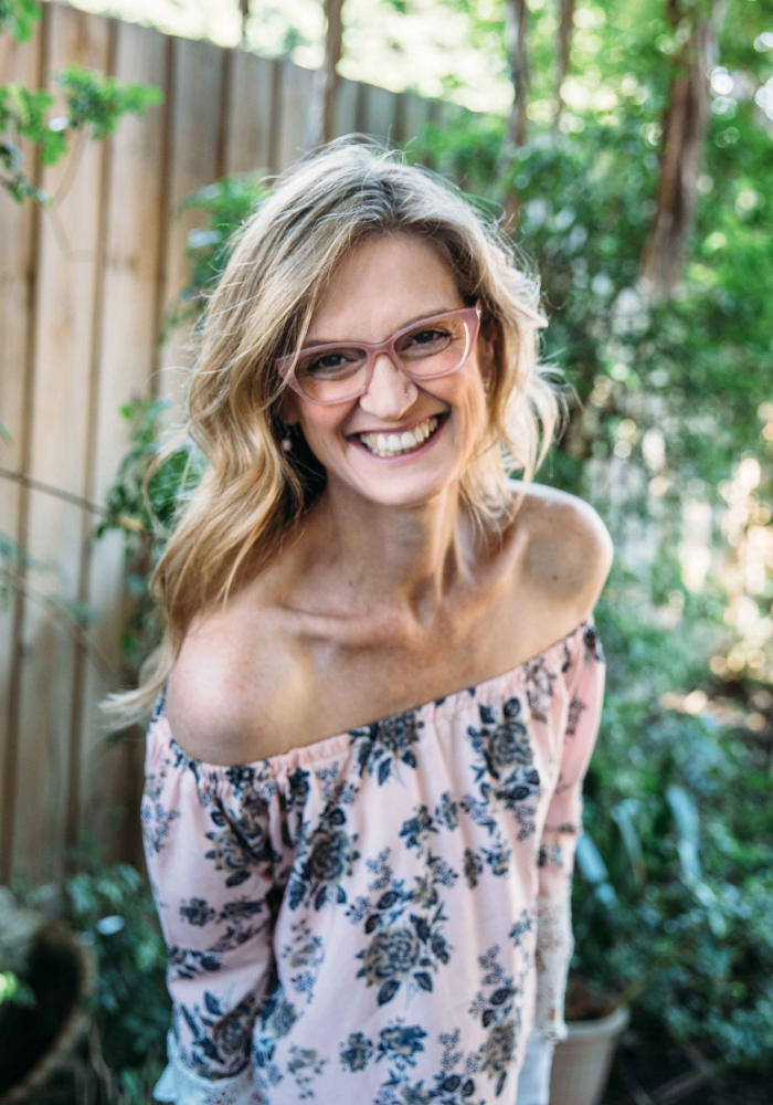 A smiling woman with blonde hair and glasses, wearing an off-shoulder floral top, standing outdoors near a wooden fence and greenery.