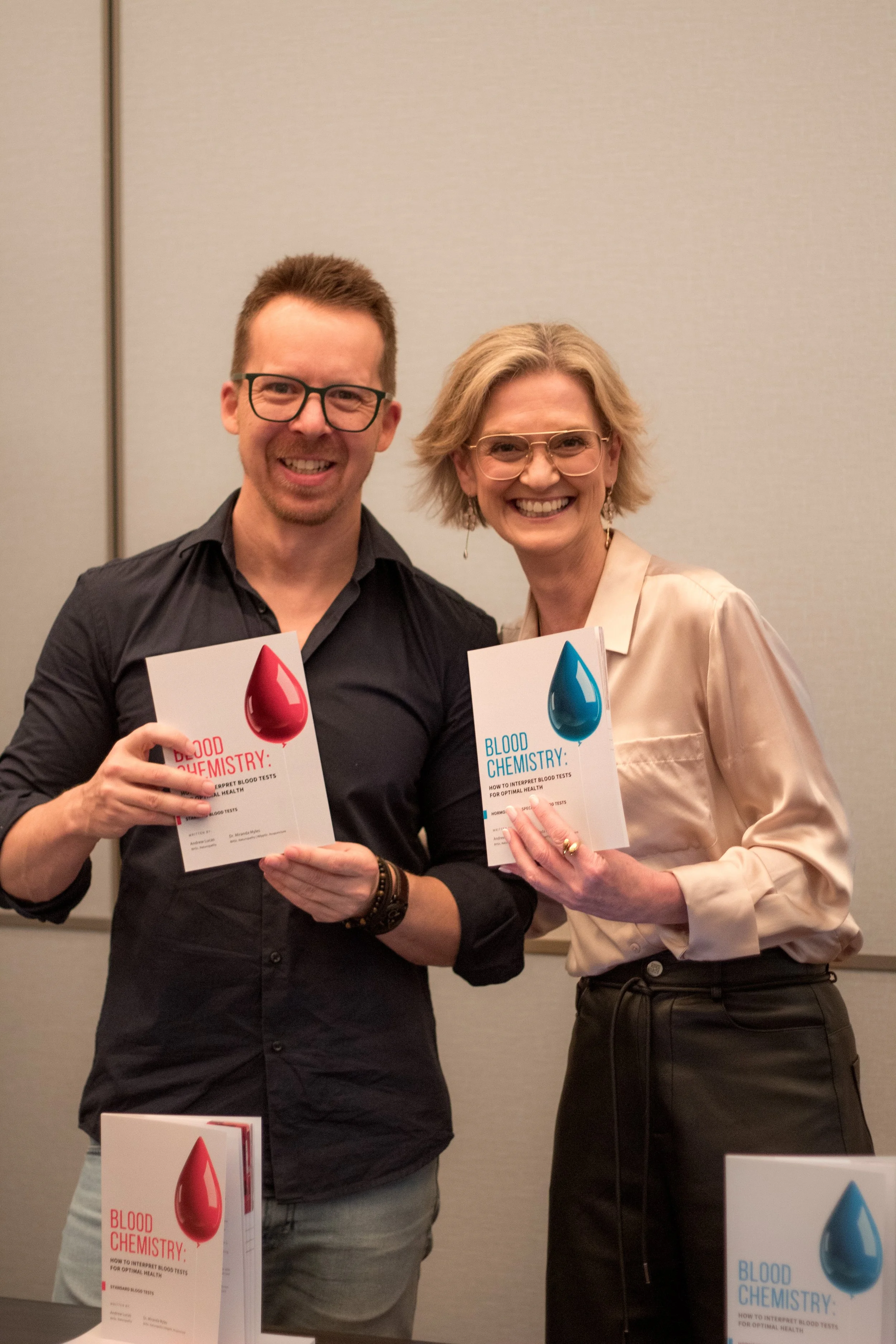Two smiling women holding books about blood chemistry at an indoor event, standing in front of a gray wall.