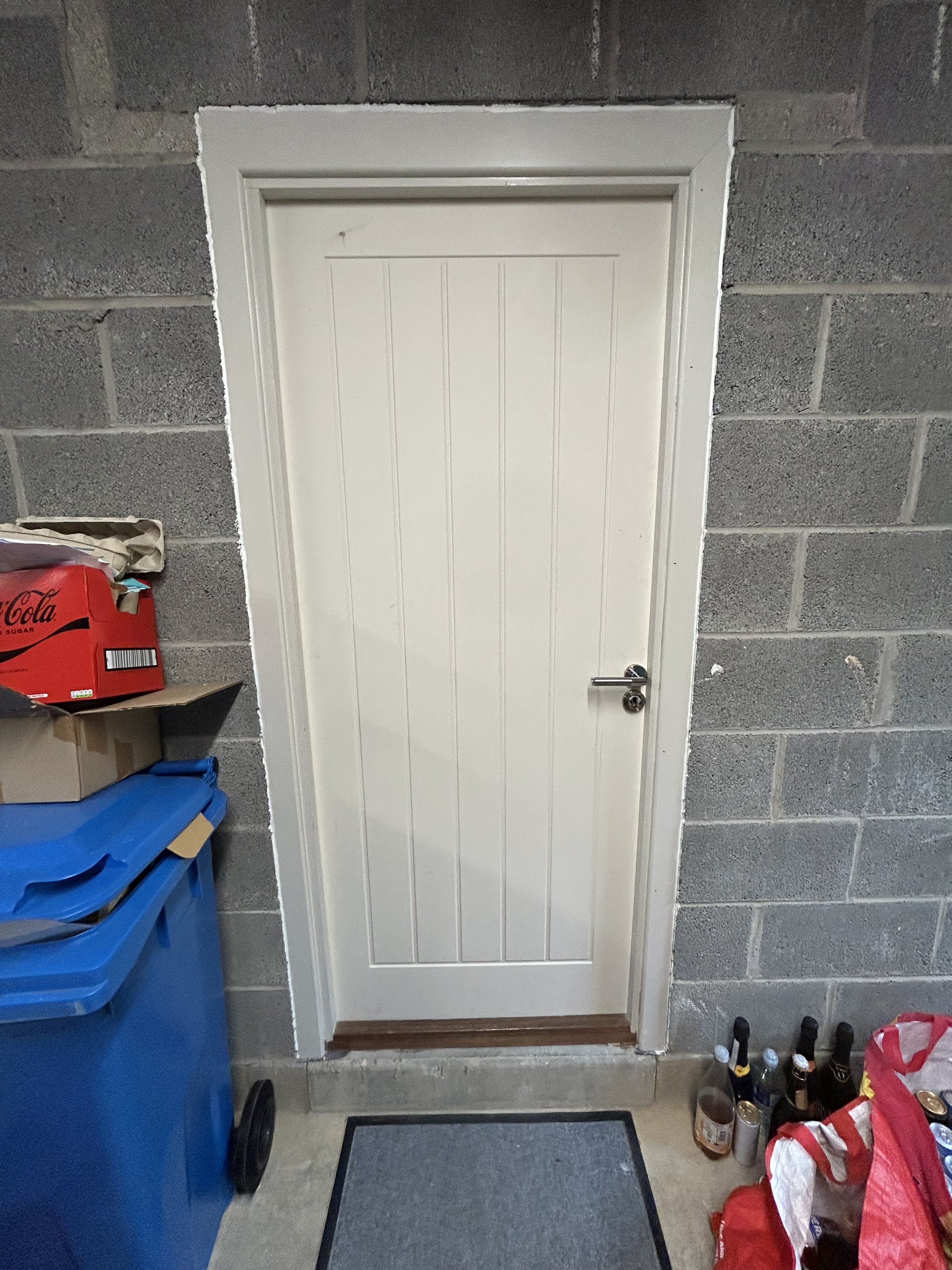 White paneled door with a silver handle in a gray cinder block wall, surrounded by storage bins, boxes, and umbrellas.