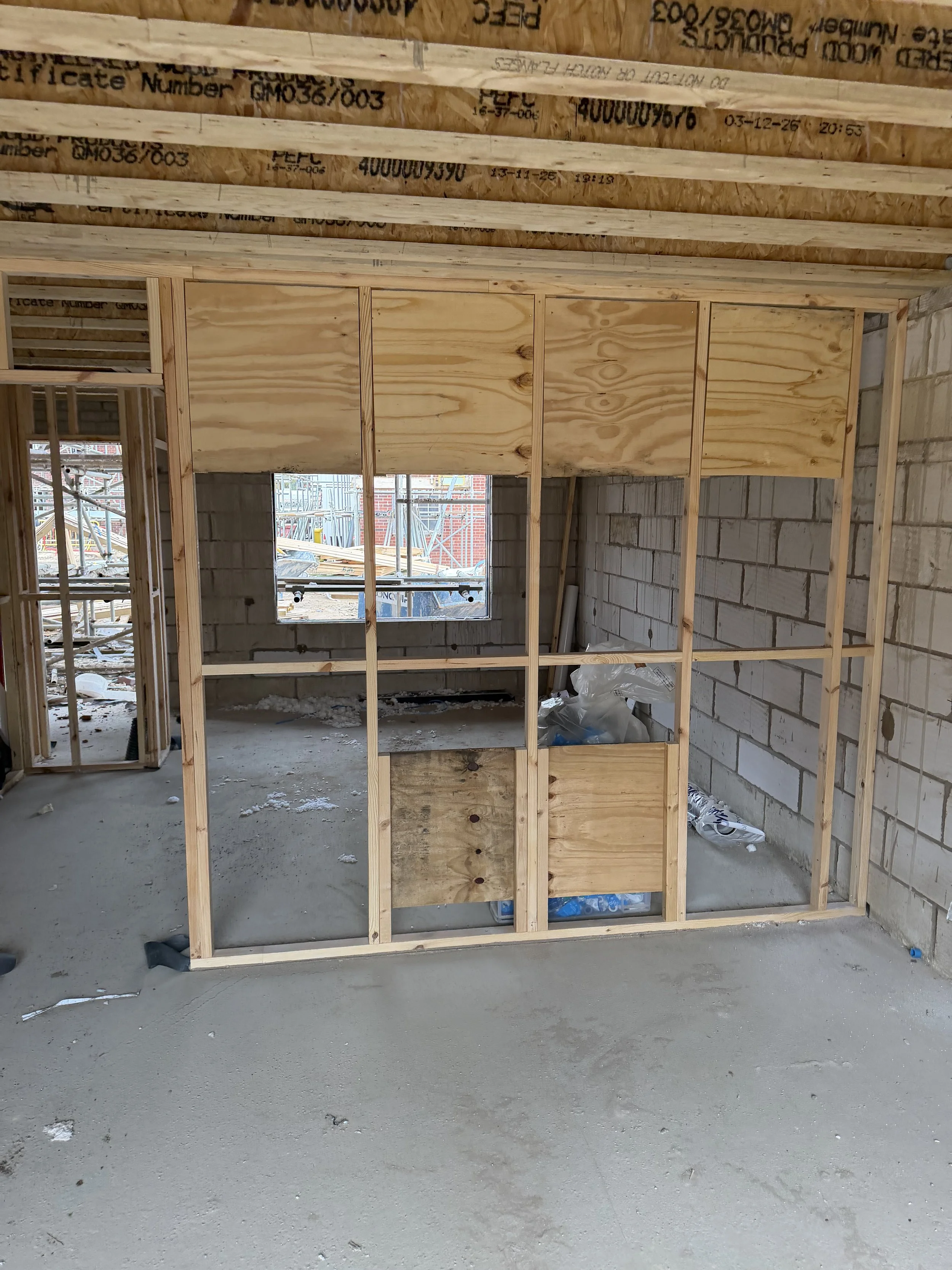 Partially framed interior of a house under construction with exposed timber studs, plywood panels, cinder block walls, windows, and concrete floor.