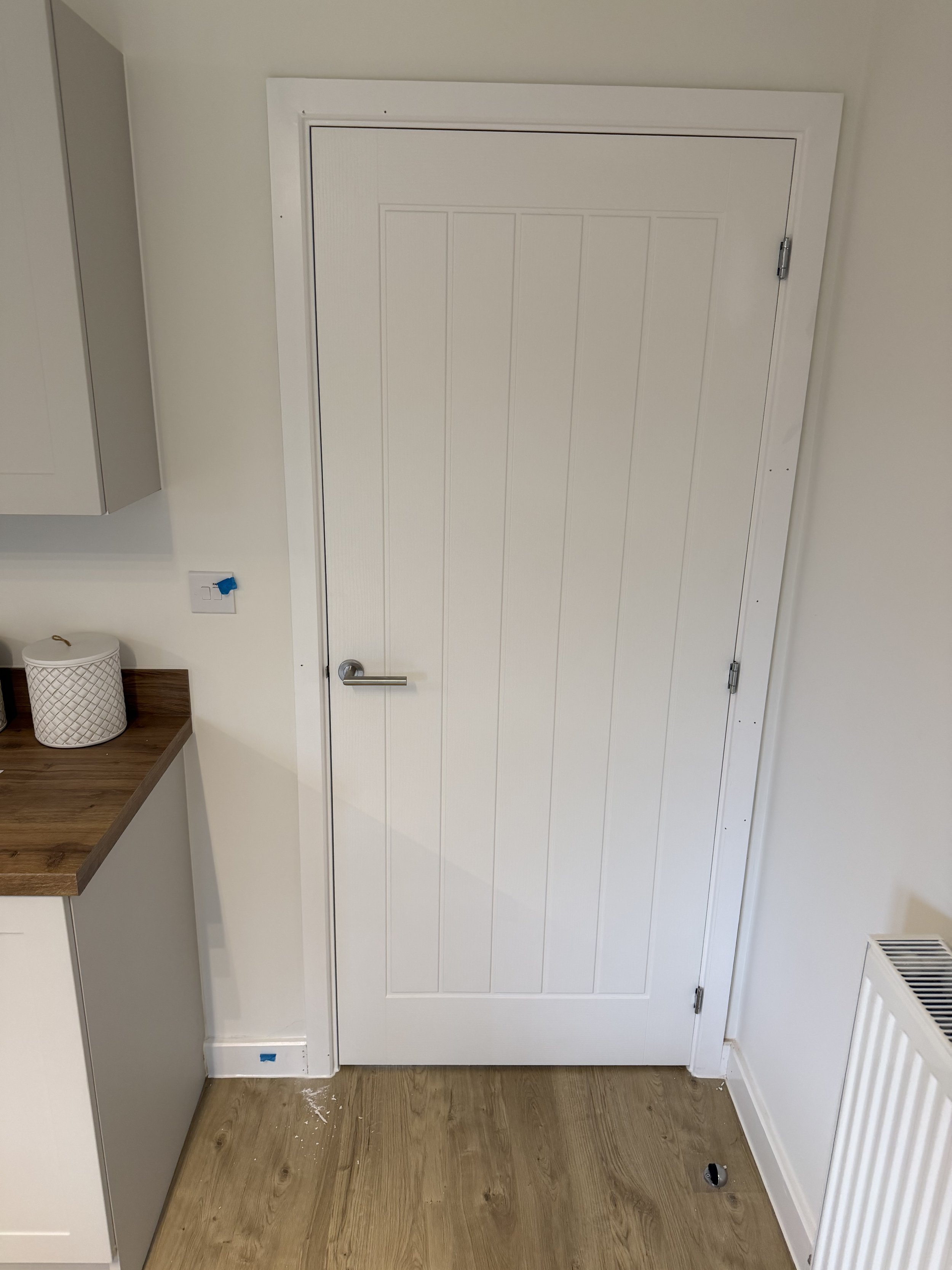 White paneled interior door with silver handle, light wood flooring, wall shelf, radiator, and woven basket
