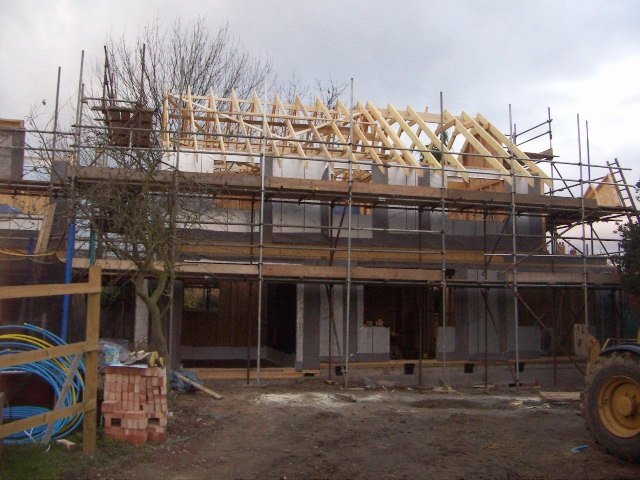 House under construction with exposed wooden roof trusses, scaffolding, and building materials on site