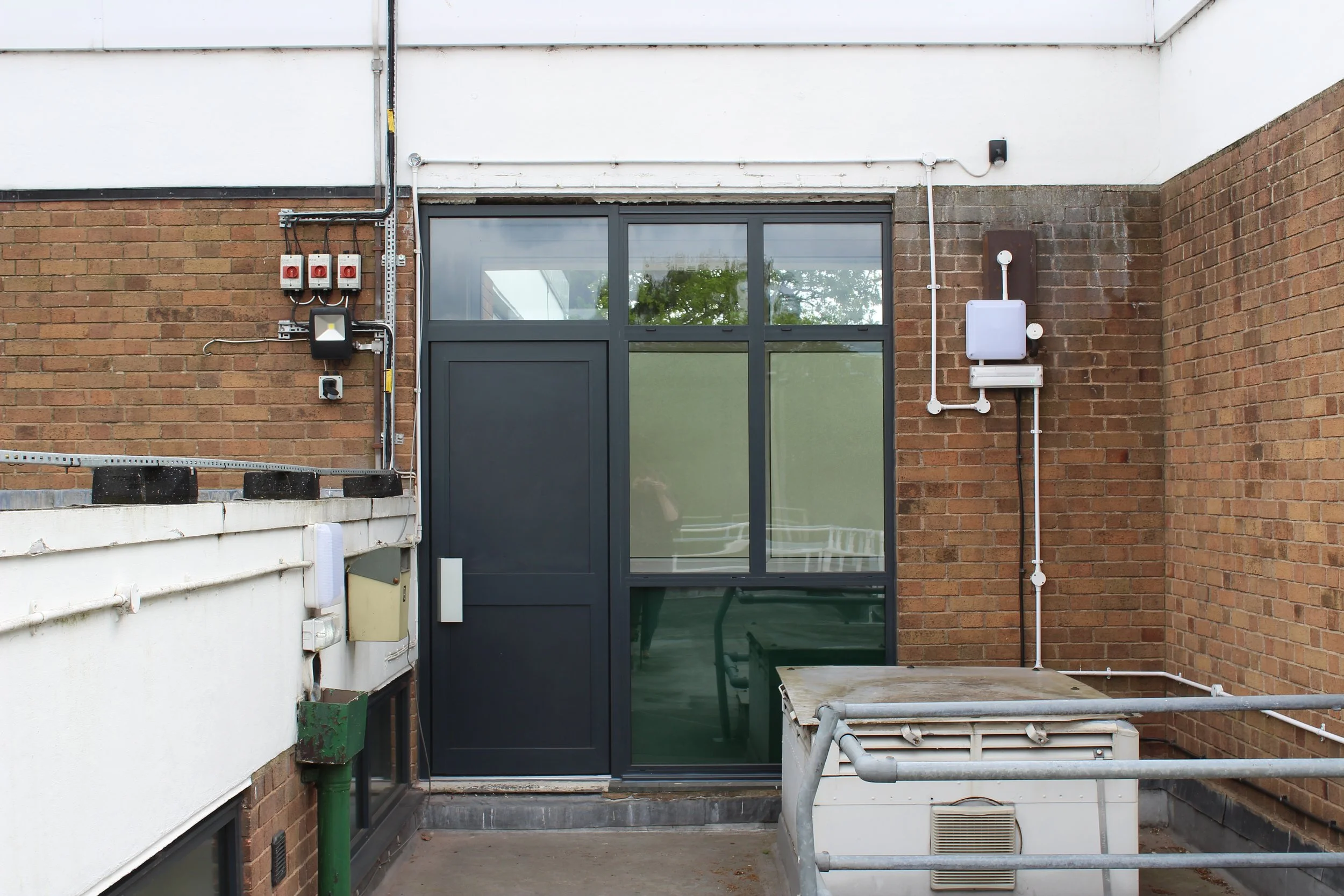 Dark-framed glass entrance door in a brick service alley with exposed electrical conduits, security lights, and outdoor HVAC equipment