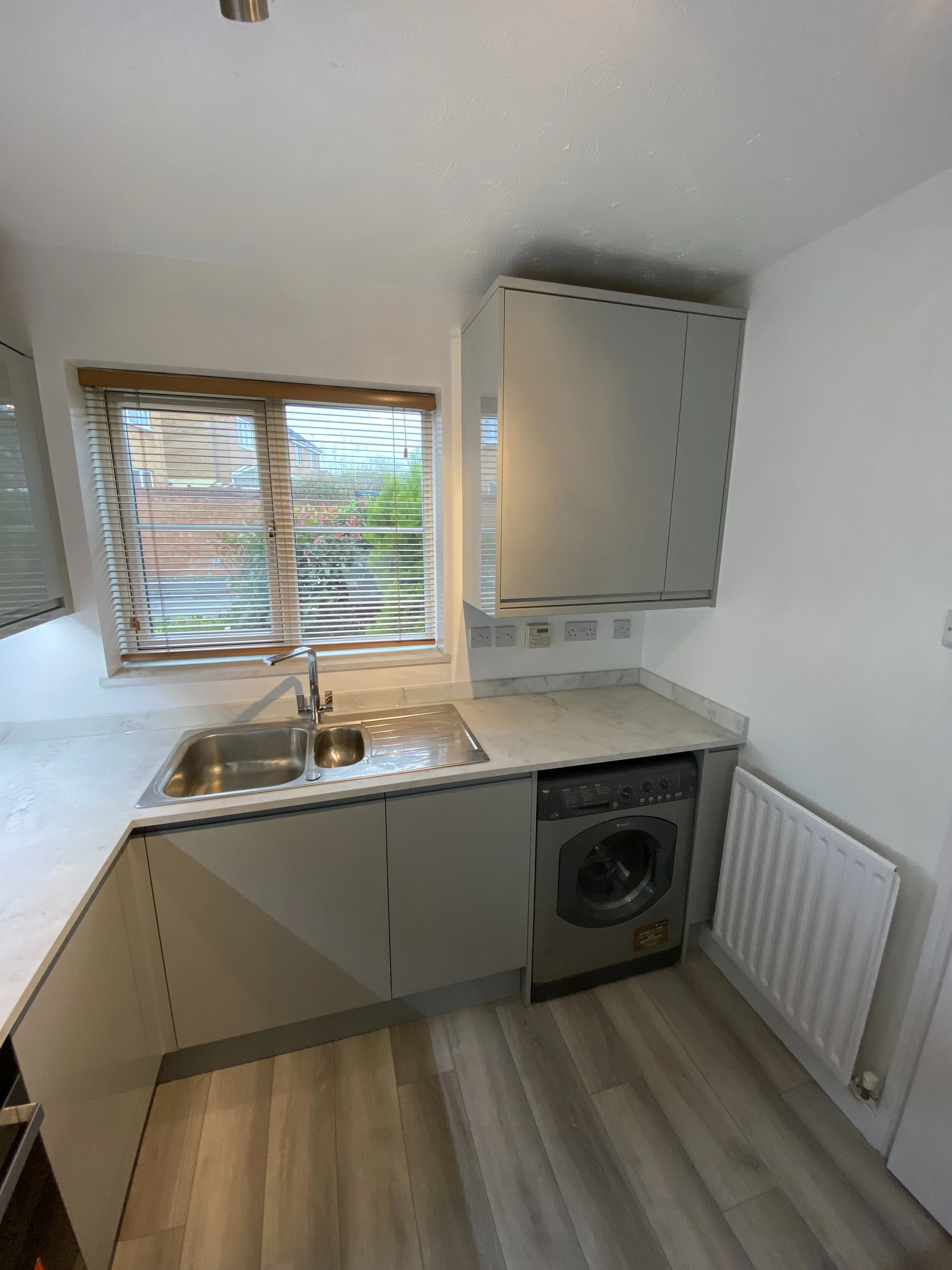 Bright modern kitchen with stainless steel sink, wood-blind window, gray cabinets, countertop, and front-loading washing machine.