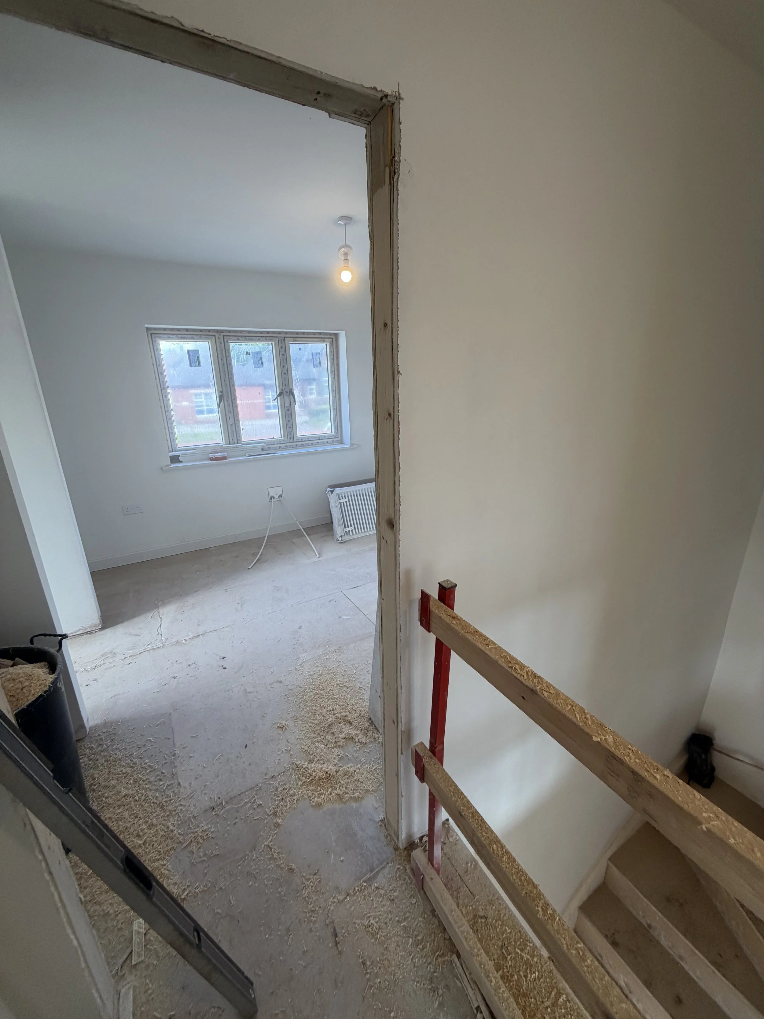Partially renovated room with exposed doorway framing, unfinished floor, new window, hanging light bulb, radiator, and construction materials.