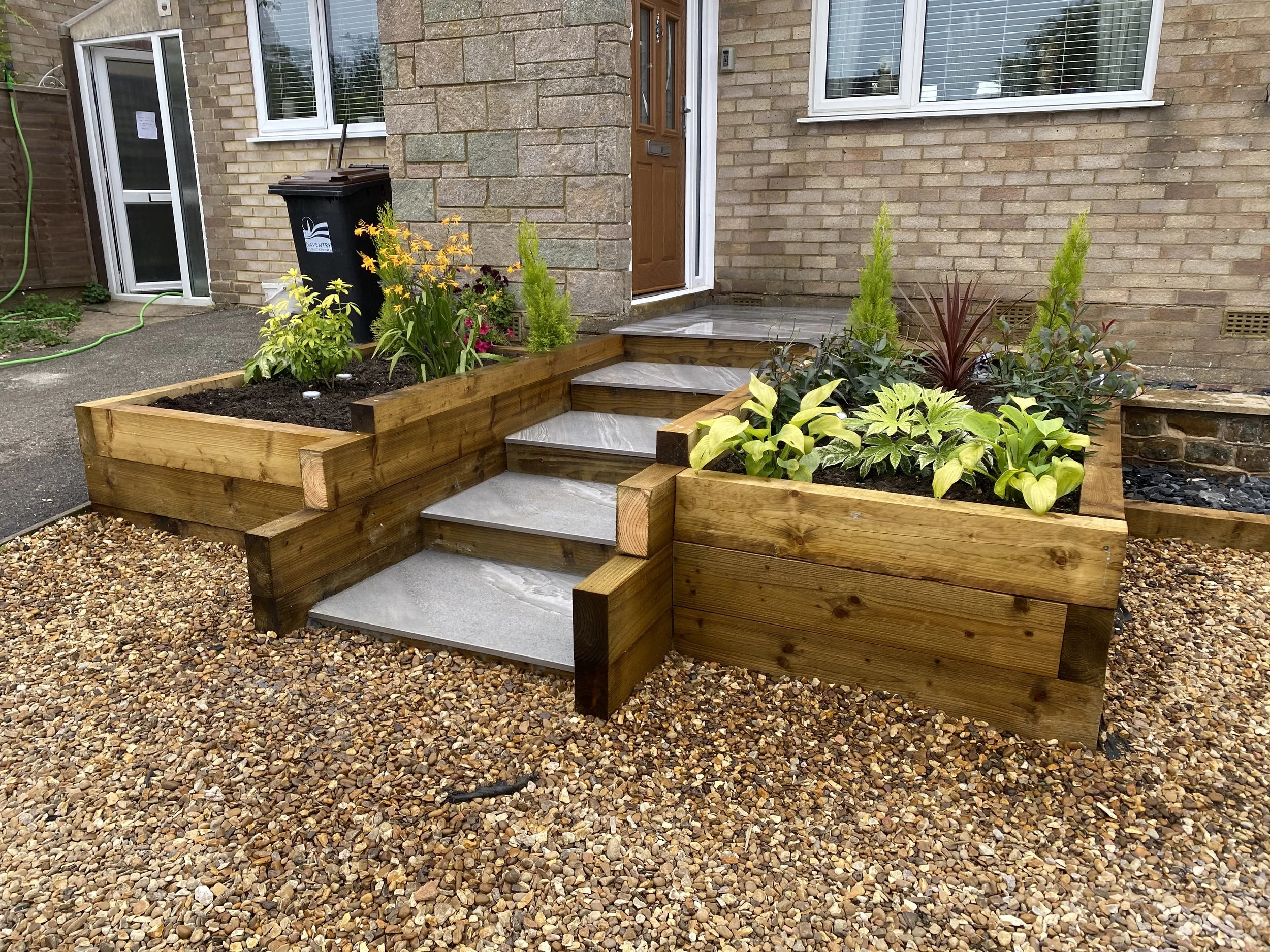 Brick house entrance with landscaped wooden raised flower beds, gray stone steps, and colorful garden plants