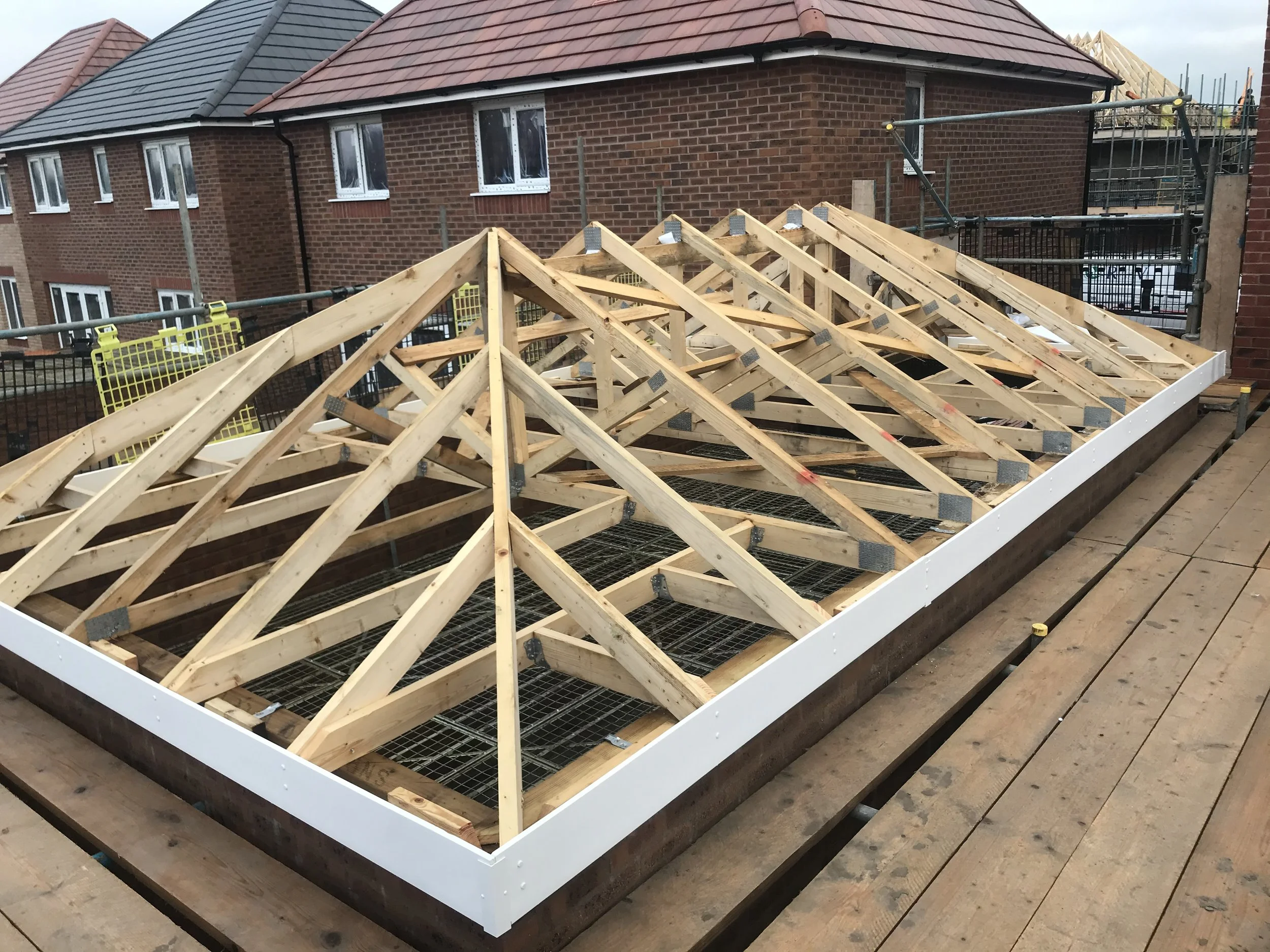 New residential roof under construction with exposed wooden trusses, scaffolding, and brick apartment buildings in the background