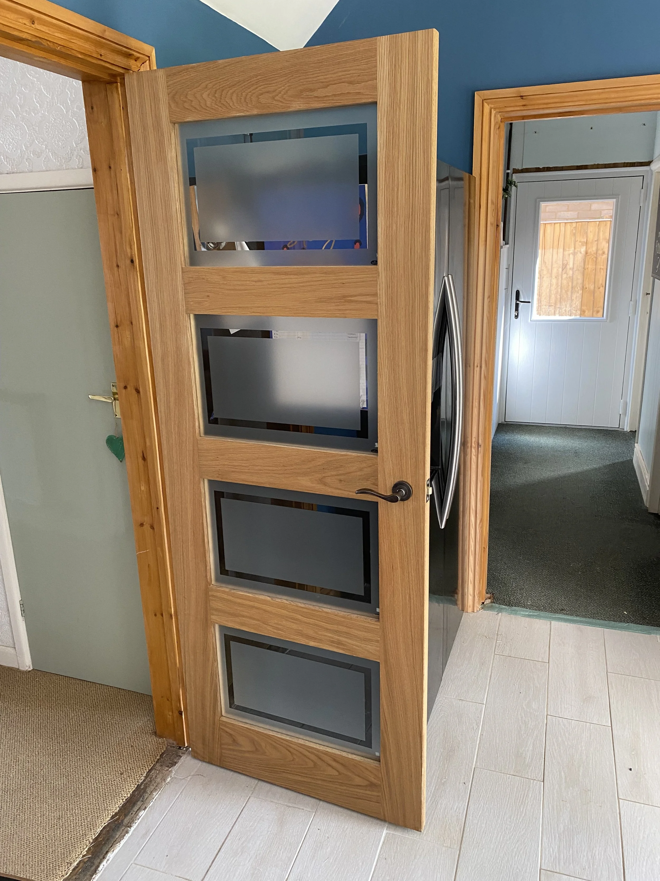 Open oak interior door with frosted glass panels and black handle in a blue-walled hallway