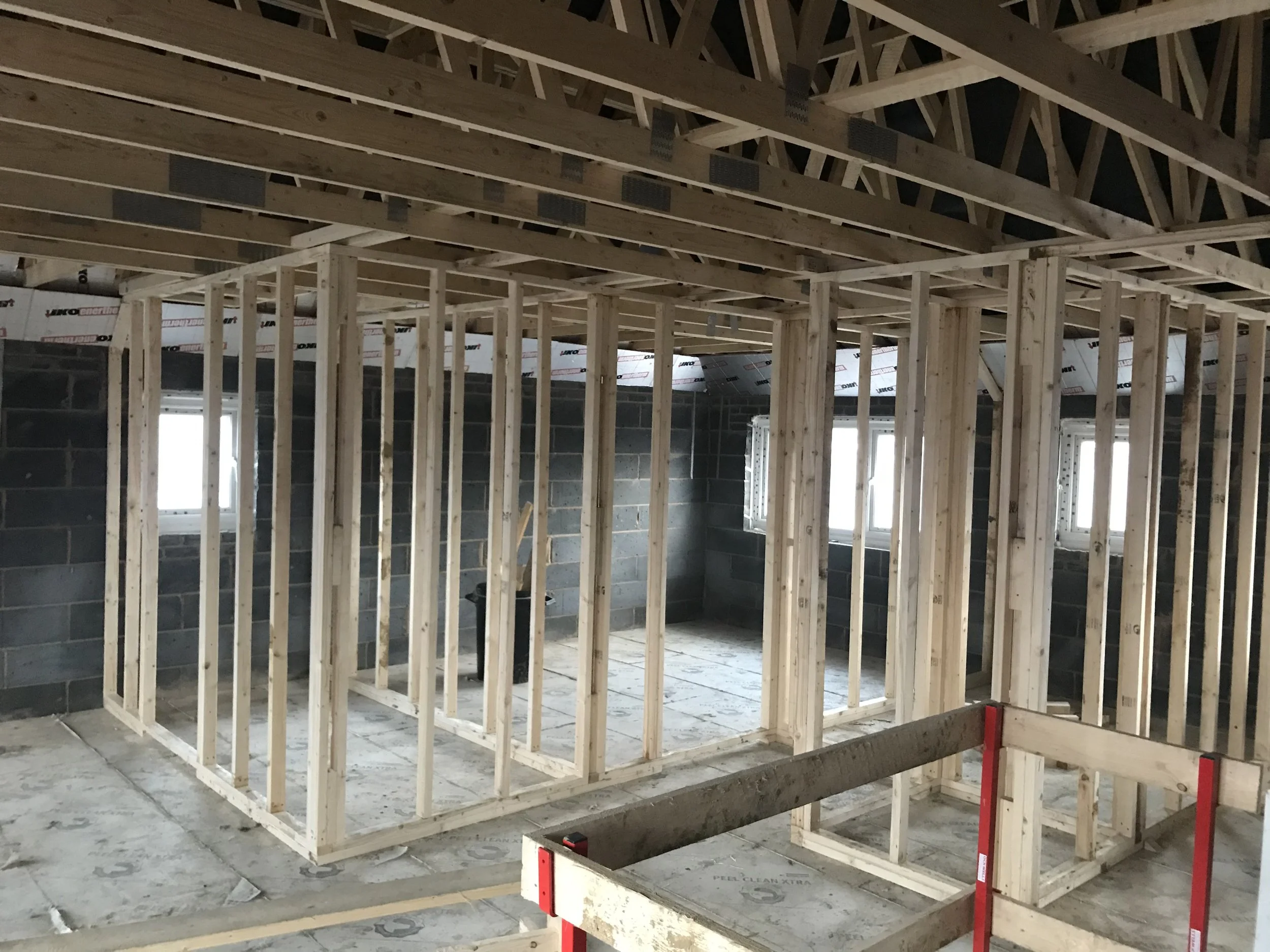 Interior of a house under construction with exposed wood framing, roof trusses, concrete block walls, windows, and a temporary work platform.