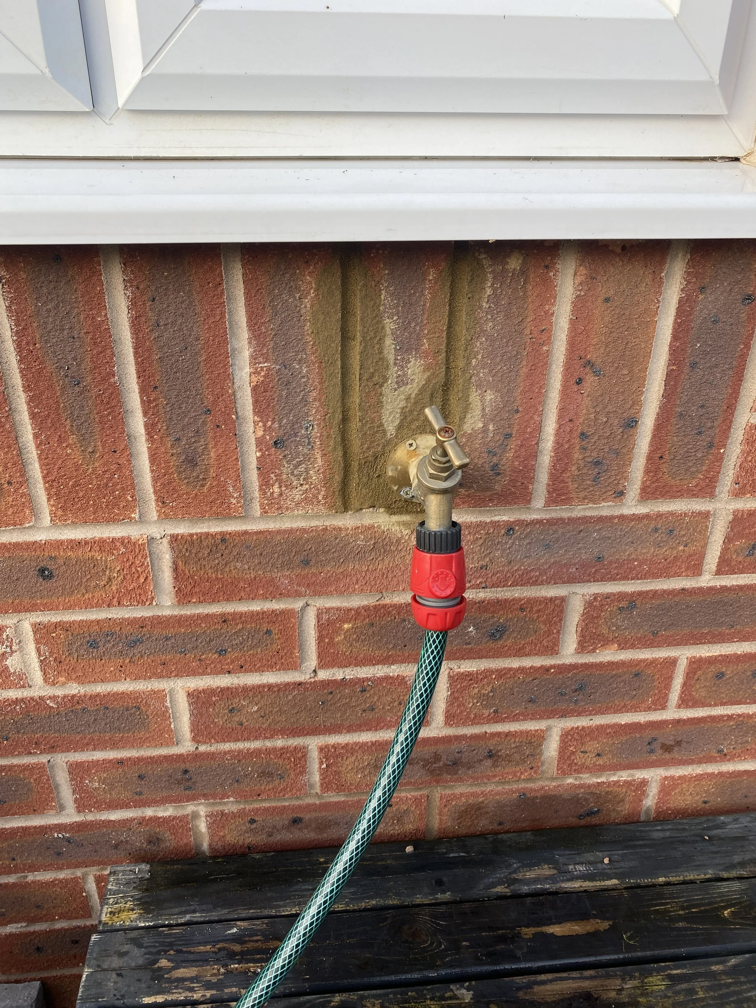Outdoor faucet with a green garden hose attached to a red connector on a brick wall beneath a white windowsill