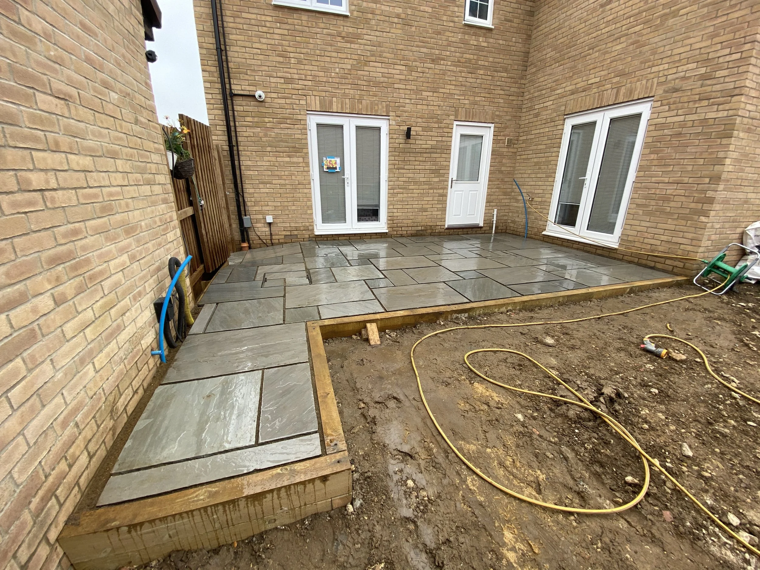 Brick-walled backyard patio with newly laid slate paving, white doors and windows, construction materials, and exposed soil.