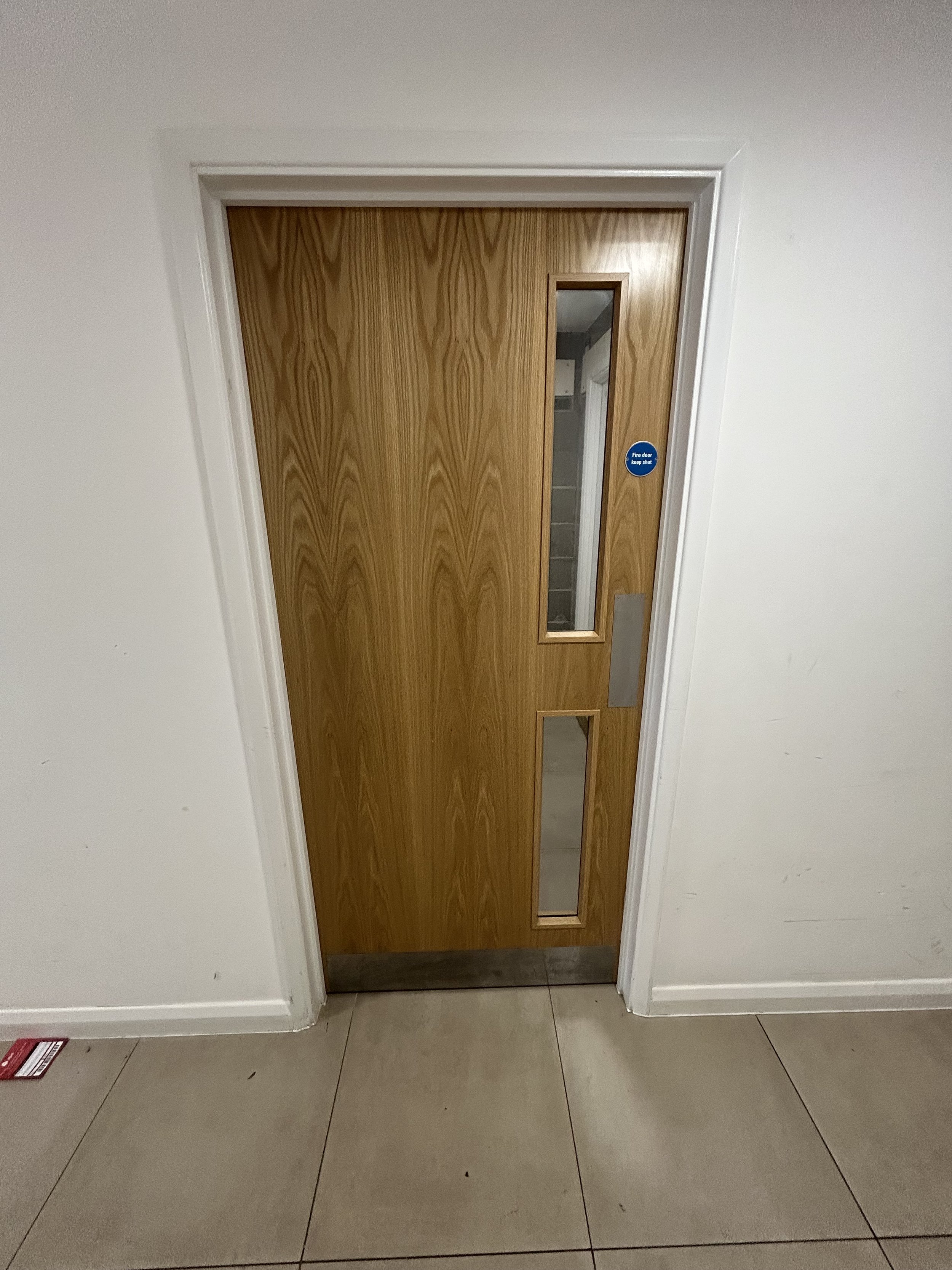 Wooden fire door with narrow vertical glass windows and a blue “Fire door keep shut” sign in a tiled hallway