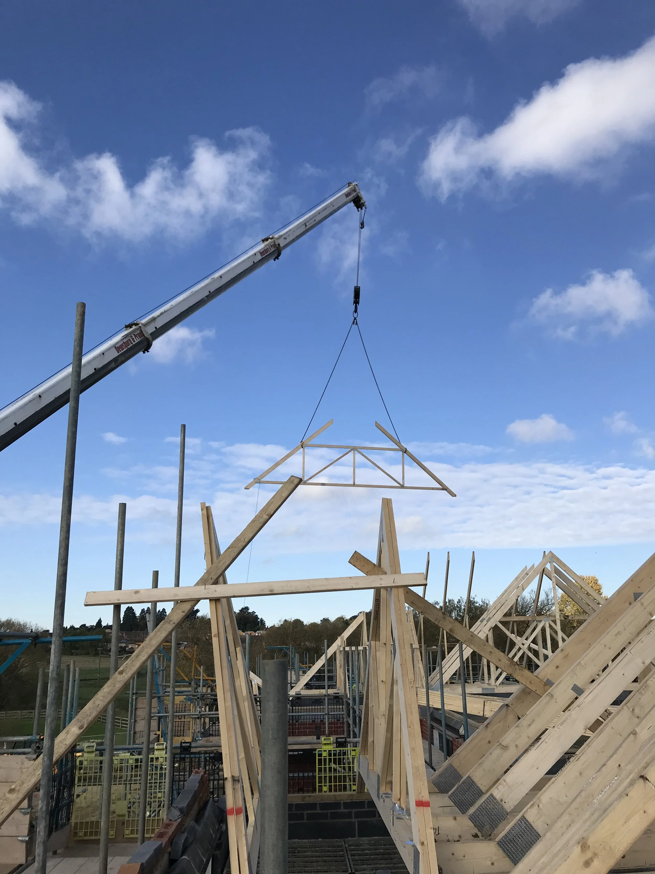 Crane lifting a wooden roof truss onto a house under construction against a blue, cloudy sky