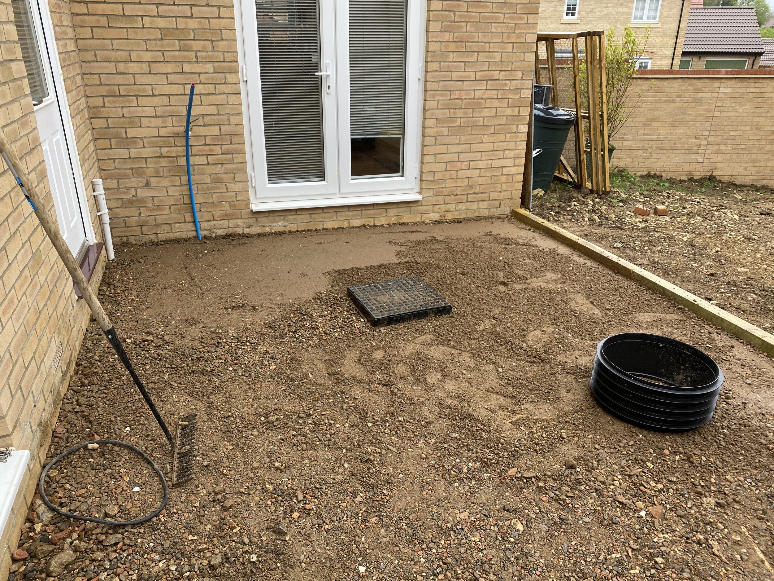 Brick house backyard under renovation, with freshly leveled gravel, drainage grate, garden borders, tools, and planters.
