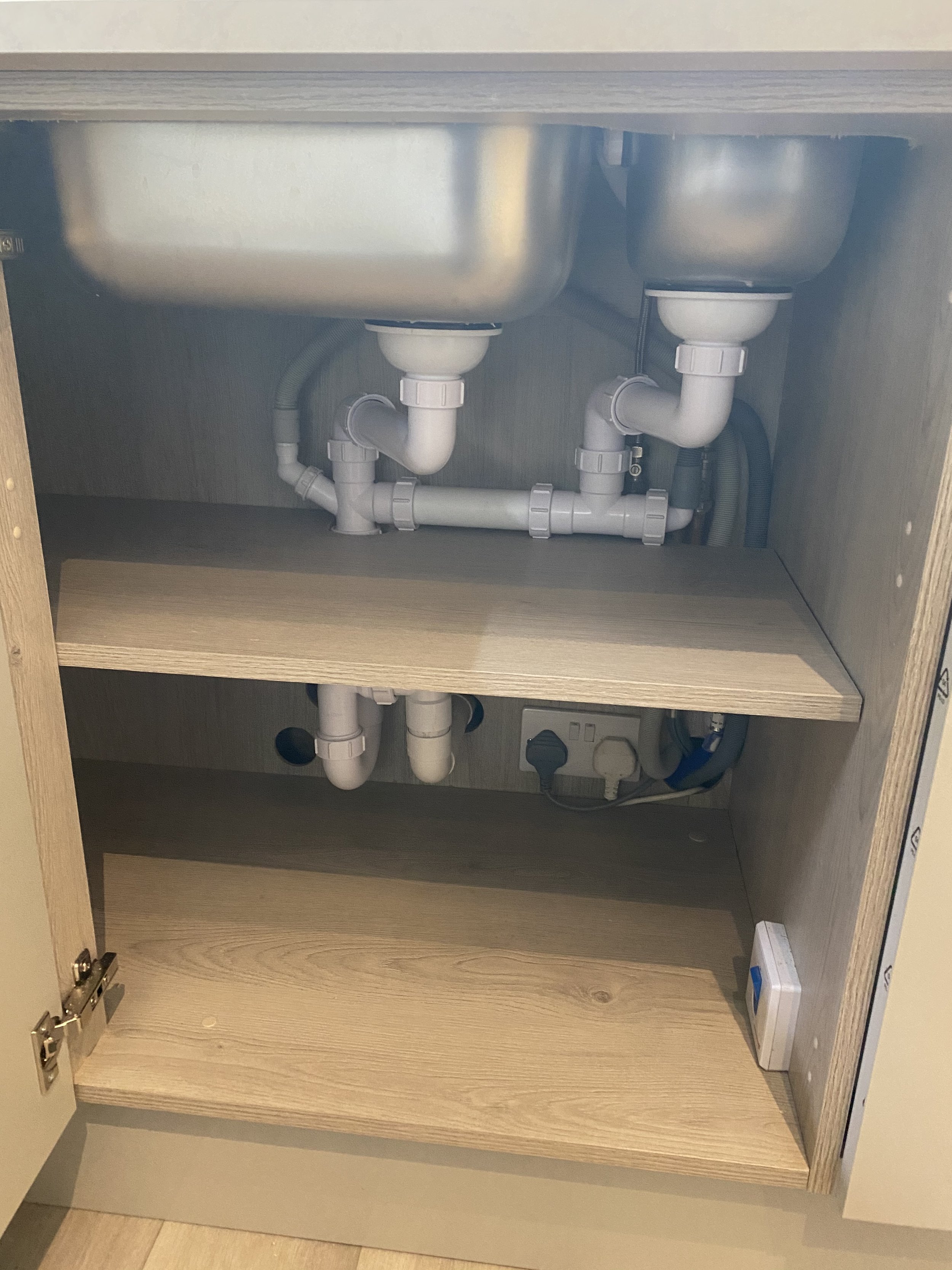 Underside of a double-basin kitchen sink showing white PVC drain pipes, P-traps, shutoff valves, and plumbing connections inside the cabinet