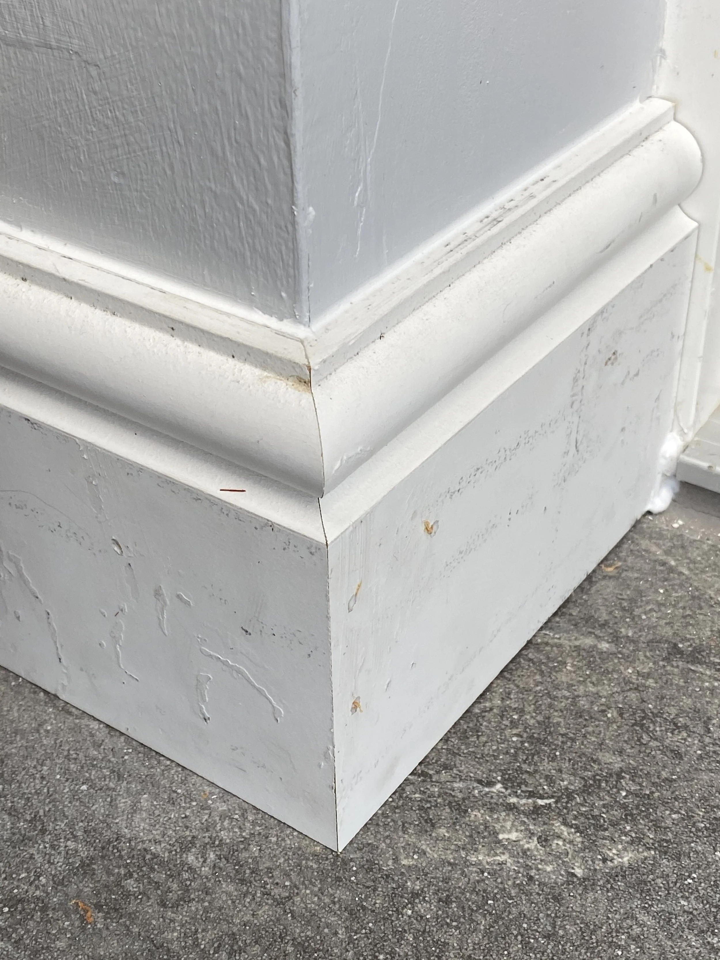 Close-up of a weathered white-painted exterior corner with layered trim, peeling paint, and asphalt pavement.
