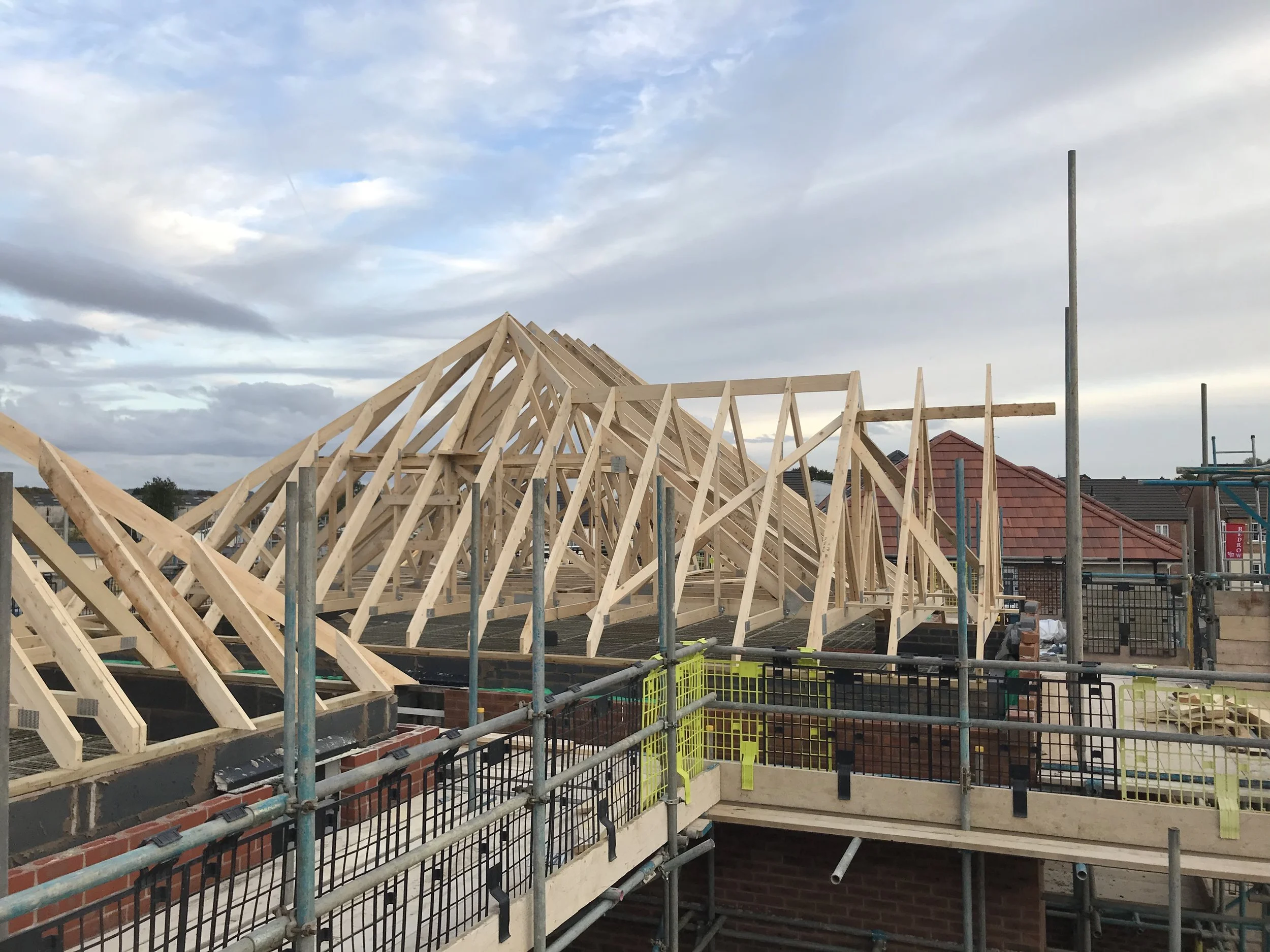 House under construction with exposed wooden roof trusses, scaffolding, and safety barriers under a cloudy sky