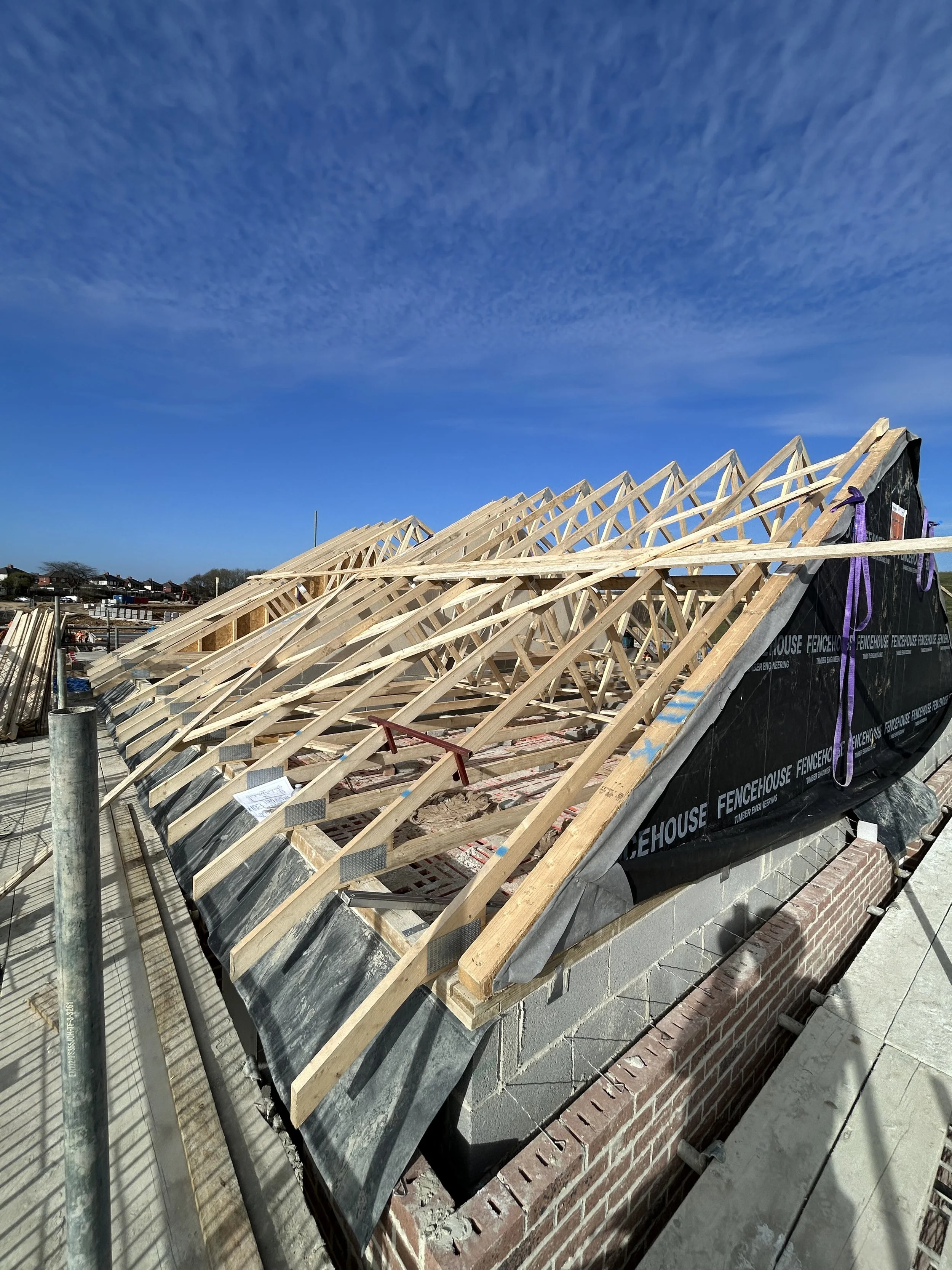 New house under construction with exposed wooden roof trusses, brickwork, and roofing membrane under a blue sky