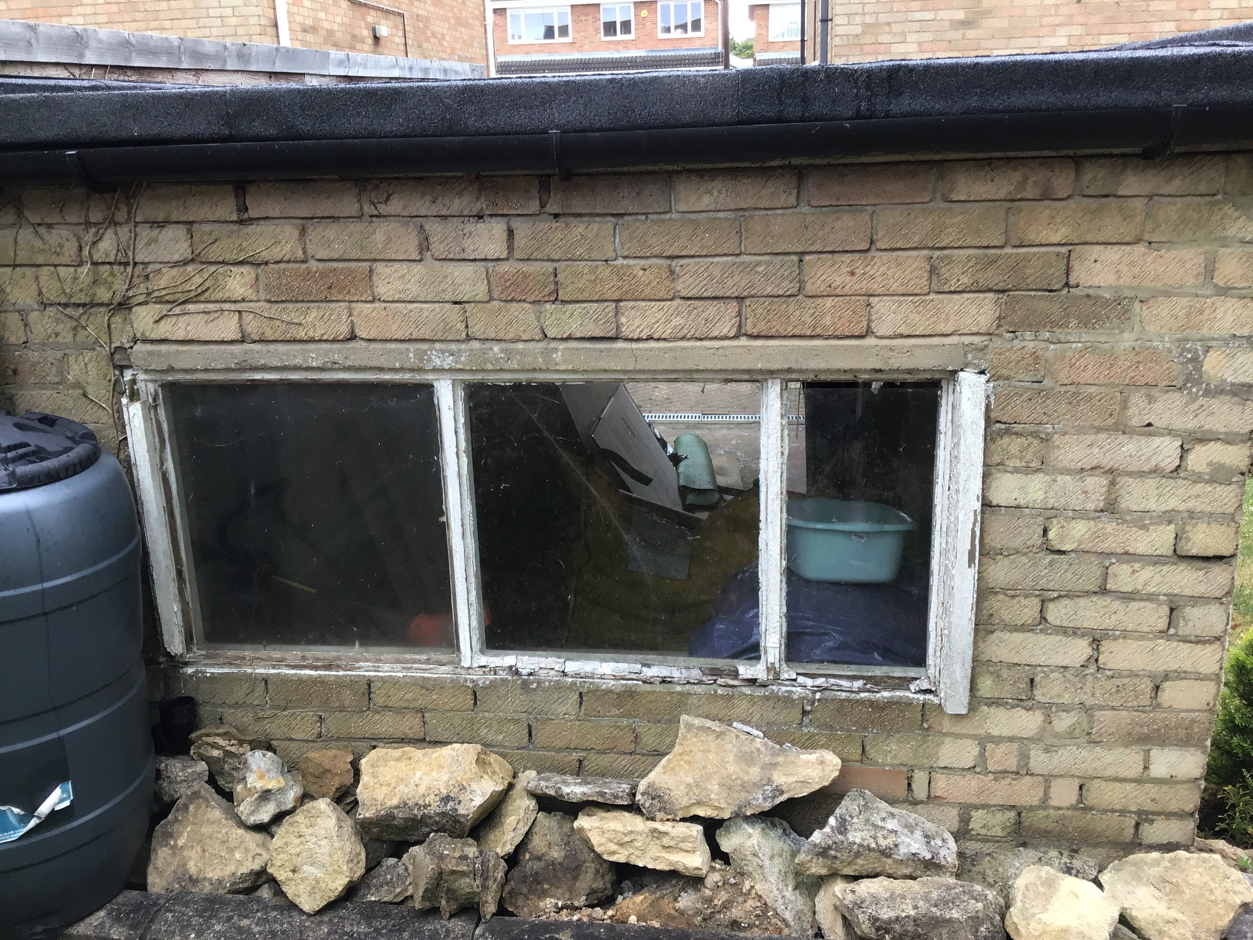Aged brick house wall with a peeling white-framed basement window, black gutter, rain barrel, and pile of landscaping stones.