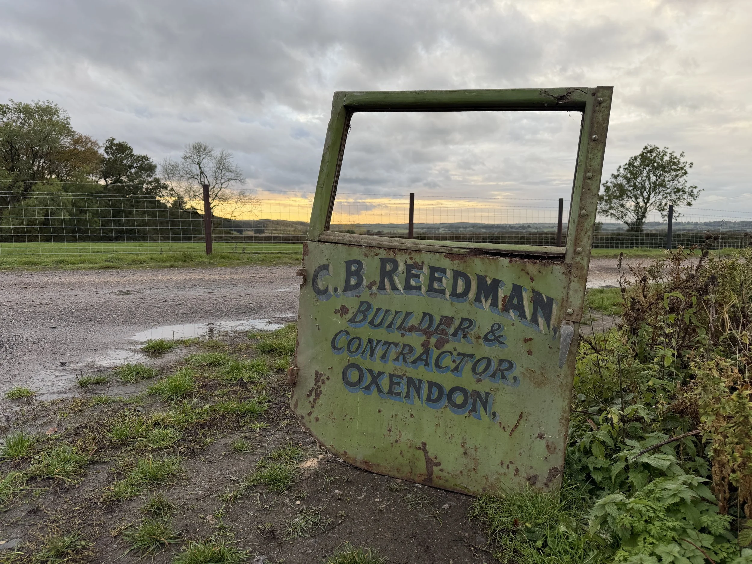 Weathered green farm sign advertising C. B. Reedman bull hire and contractor services beside a wire-fenced field at sunset.