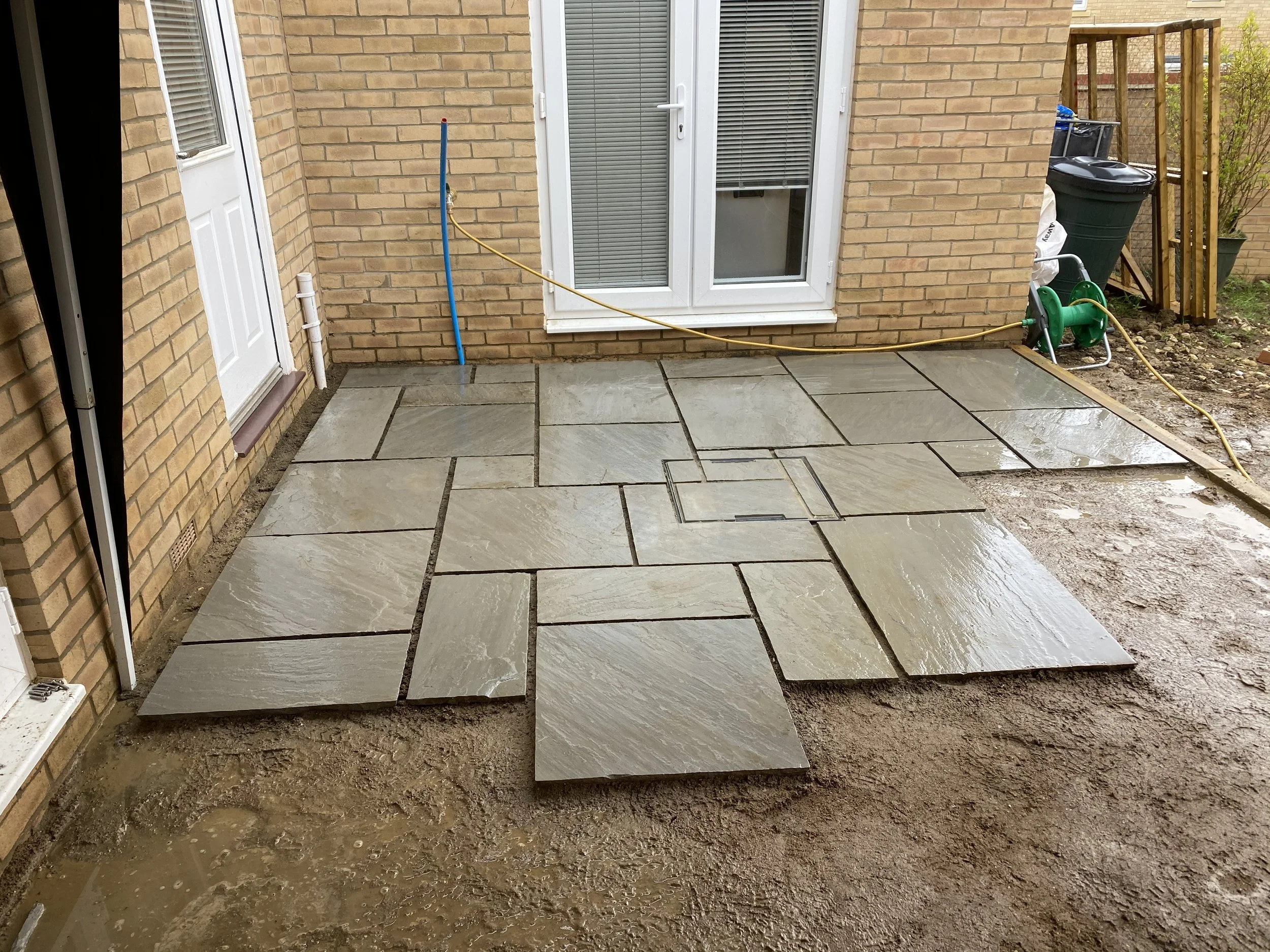 Sandstone patio being installed beside a brick house, with a garden hose, watering reel, trash bin, and shovel visible.