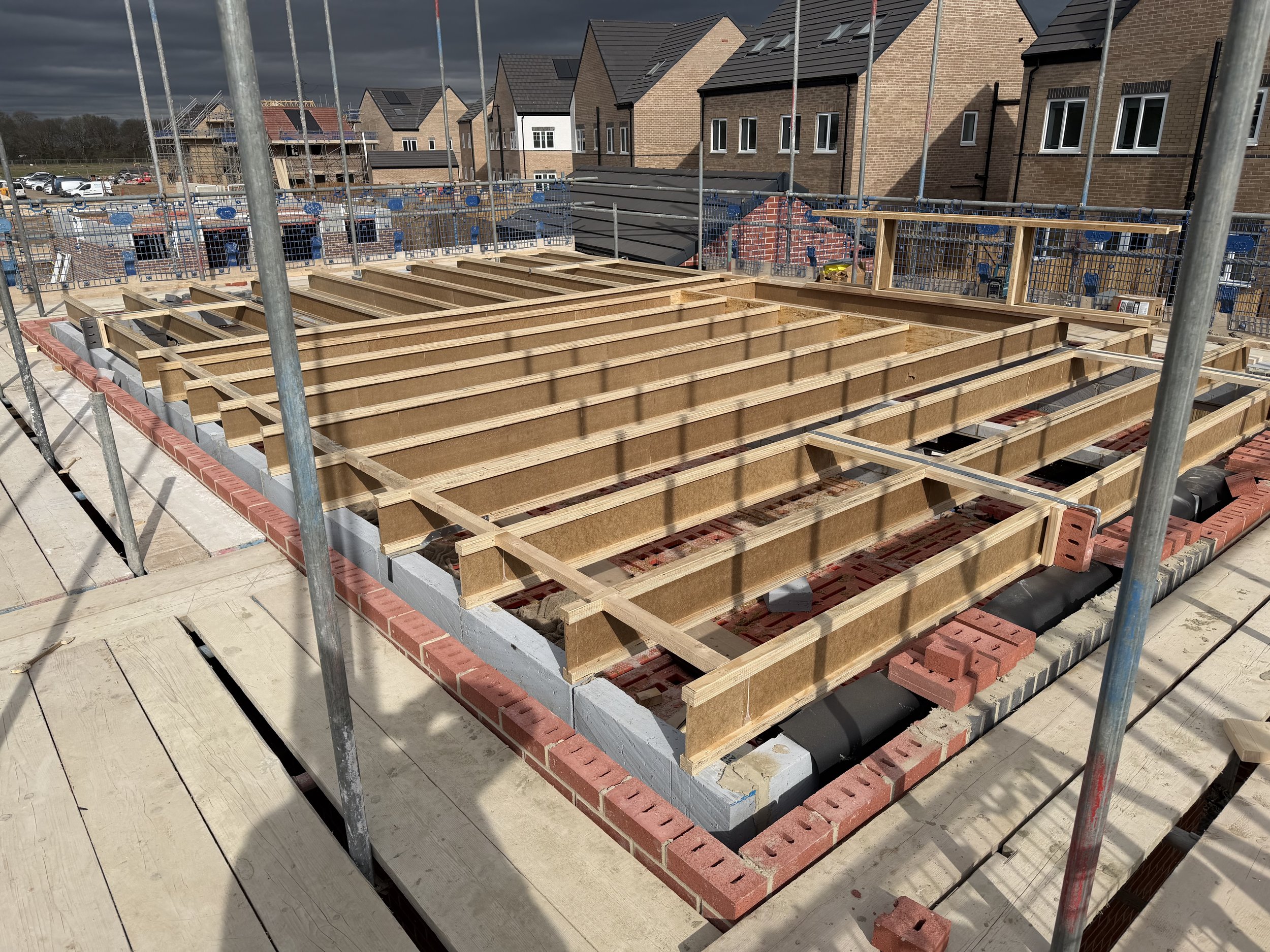 New residential construction site showing timber floor joists, brickwork, scaffolding, and completed brick houses under dark clouds