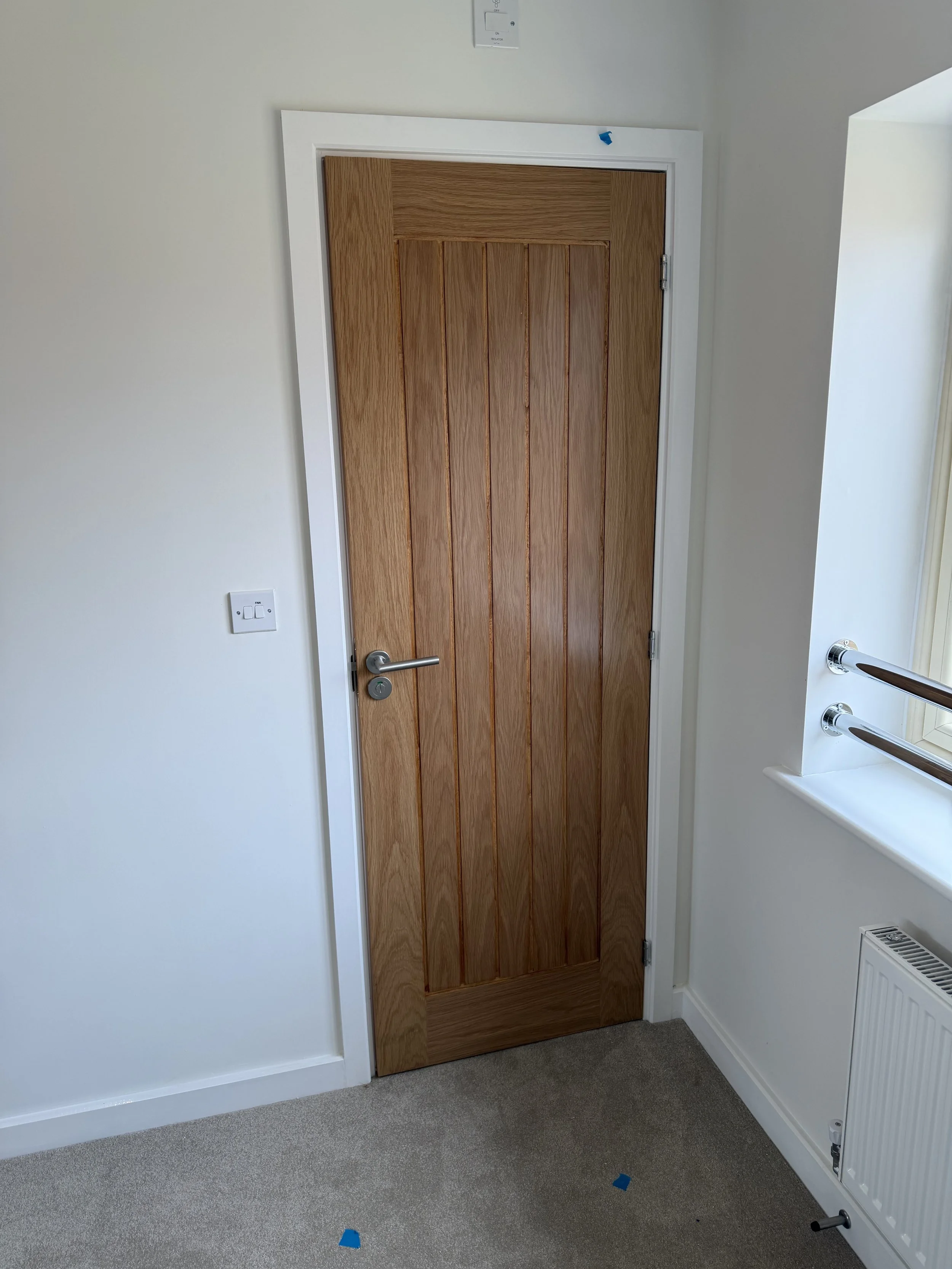 Oak paneled interior door with silver handle in a carpeted room, beside a wall-mounted radiator and grab rails.