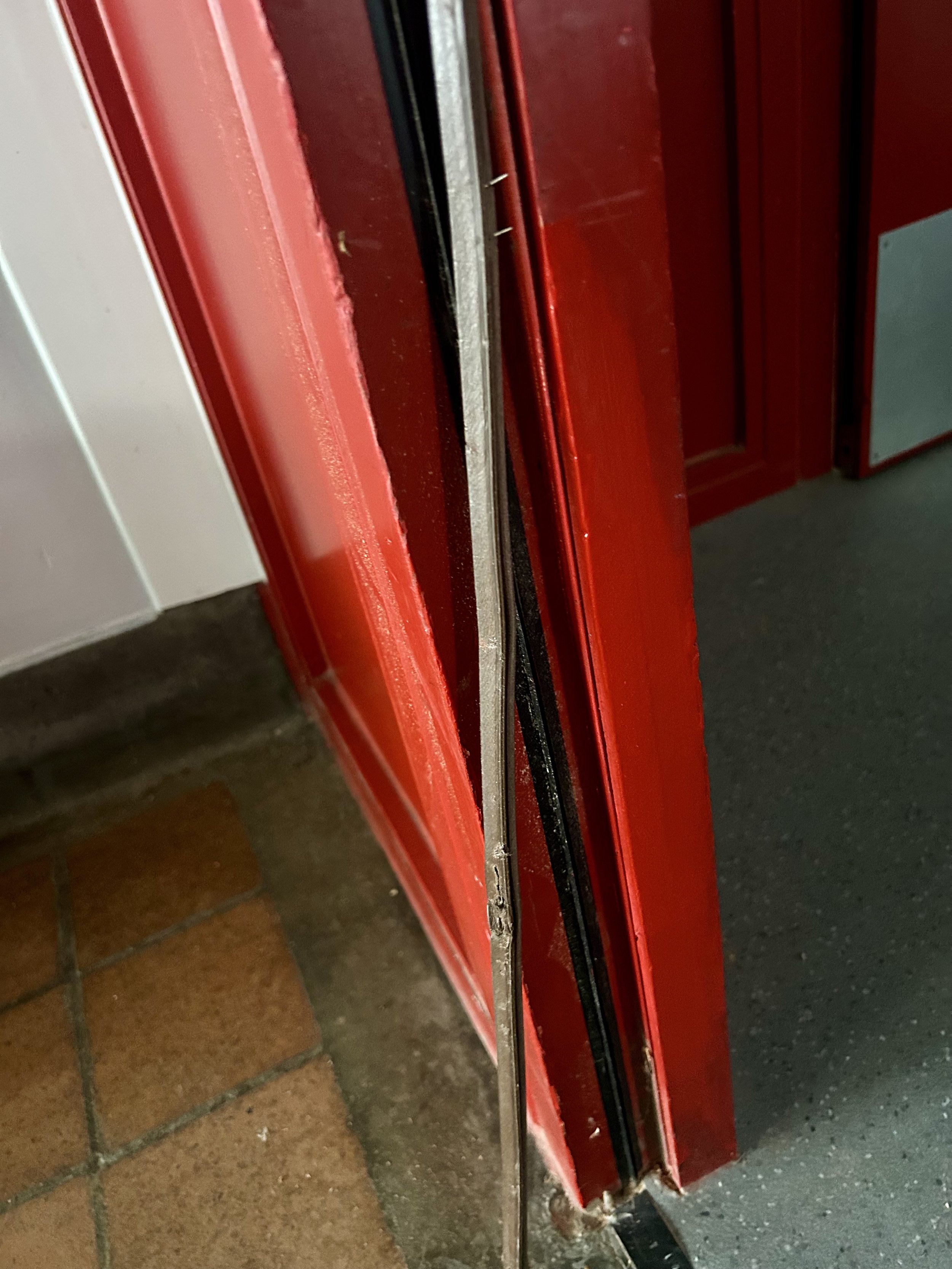 Close-up of a damaged red metal door frame with exposed weatherstripping and a bent metal strip.