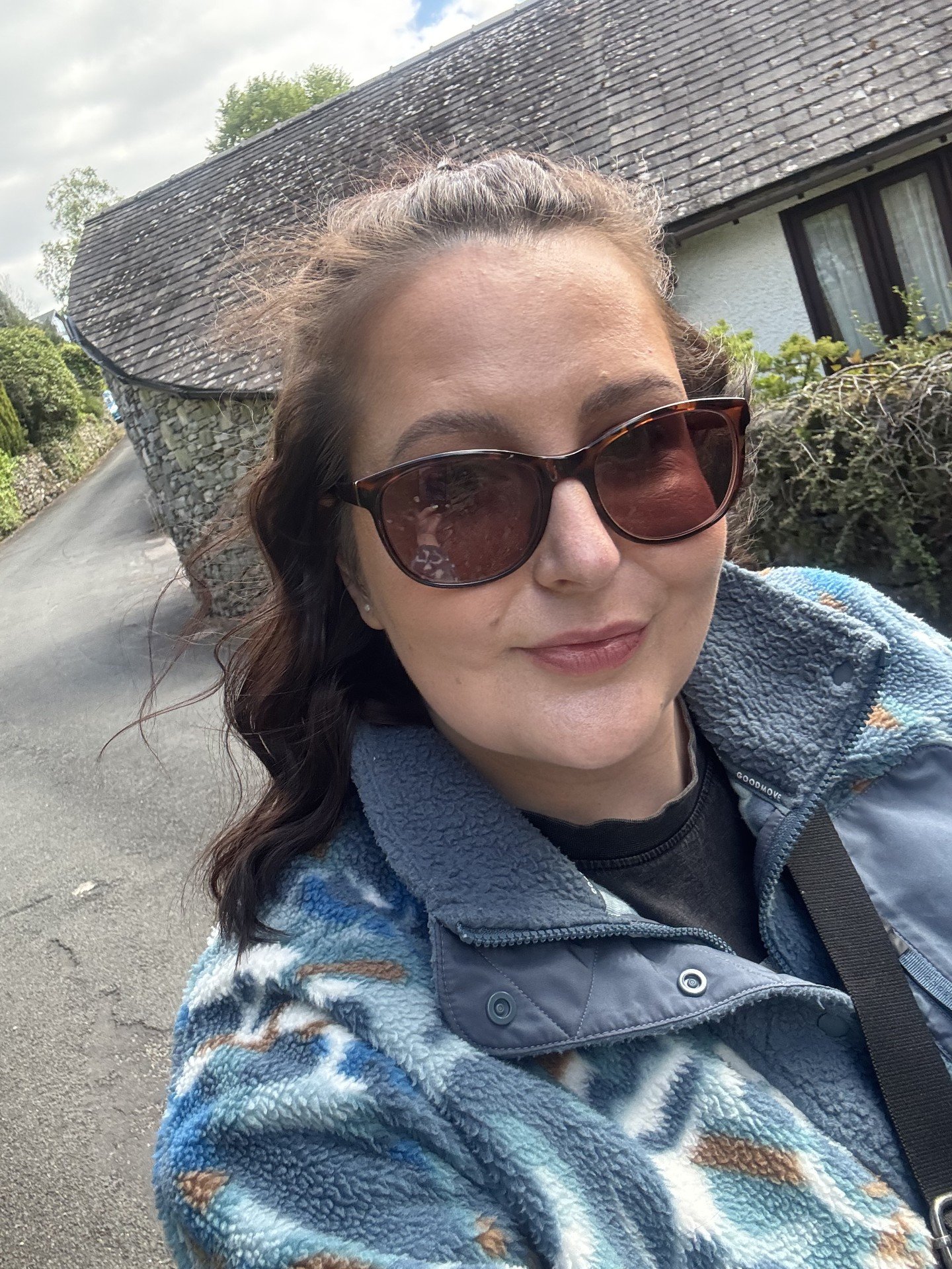 A woman with dark wavy hair wearing sunglasses, a patterned fleece jacket, and a black shirt, taking a selfie outdoors in front of a house with a stone wall and gray roof.