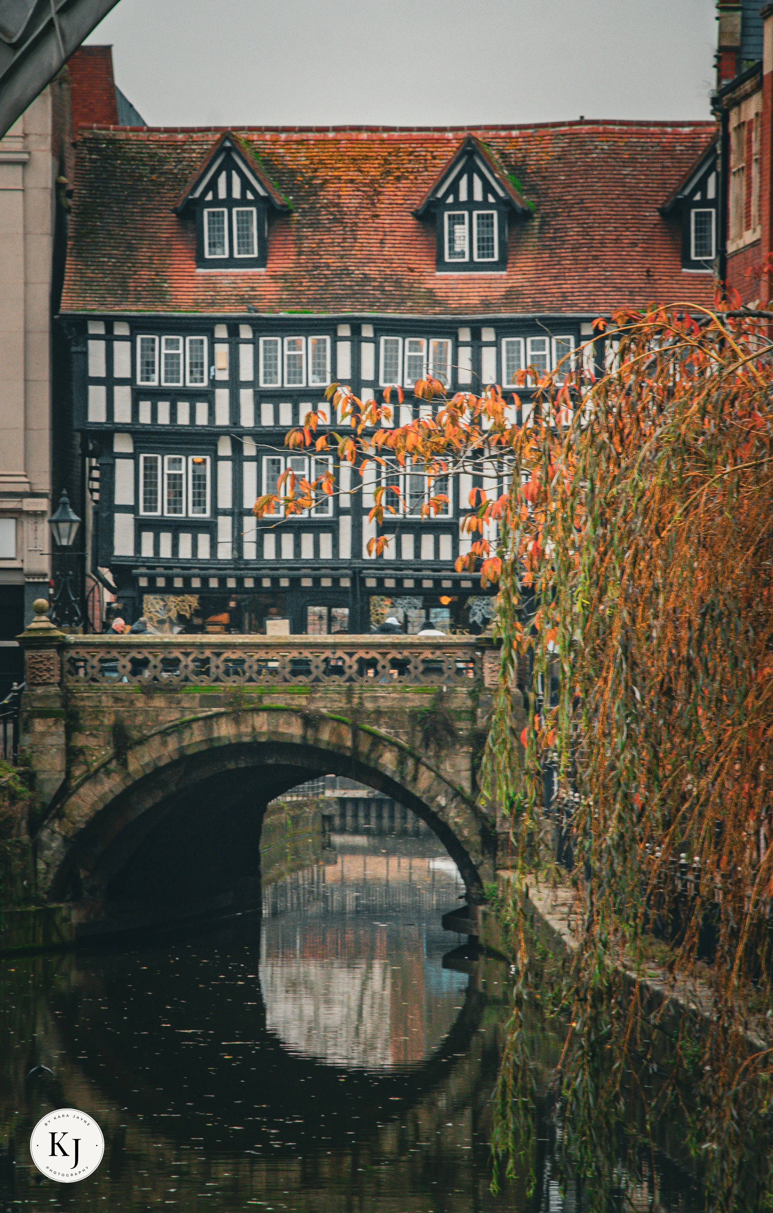 A historic black and white timber-framed building with a red tiled roof arches over a canal with an stone bridge, autumn leaves, and water reflections.