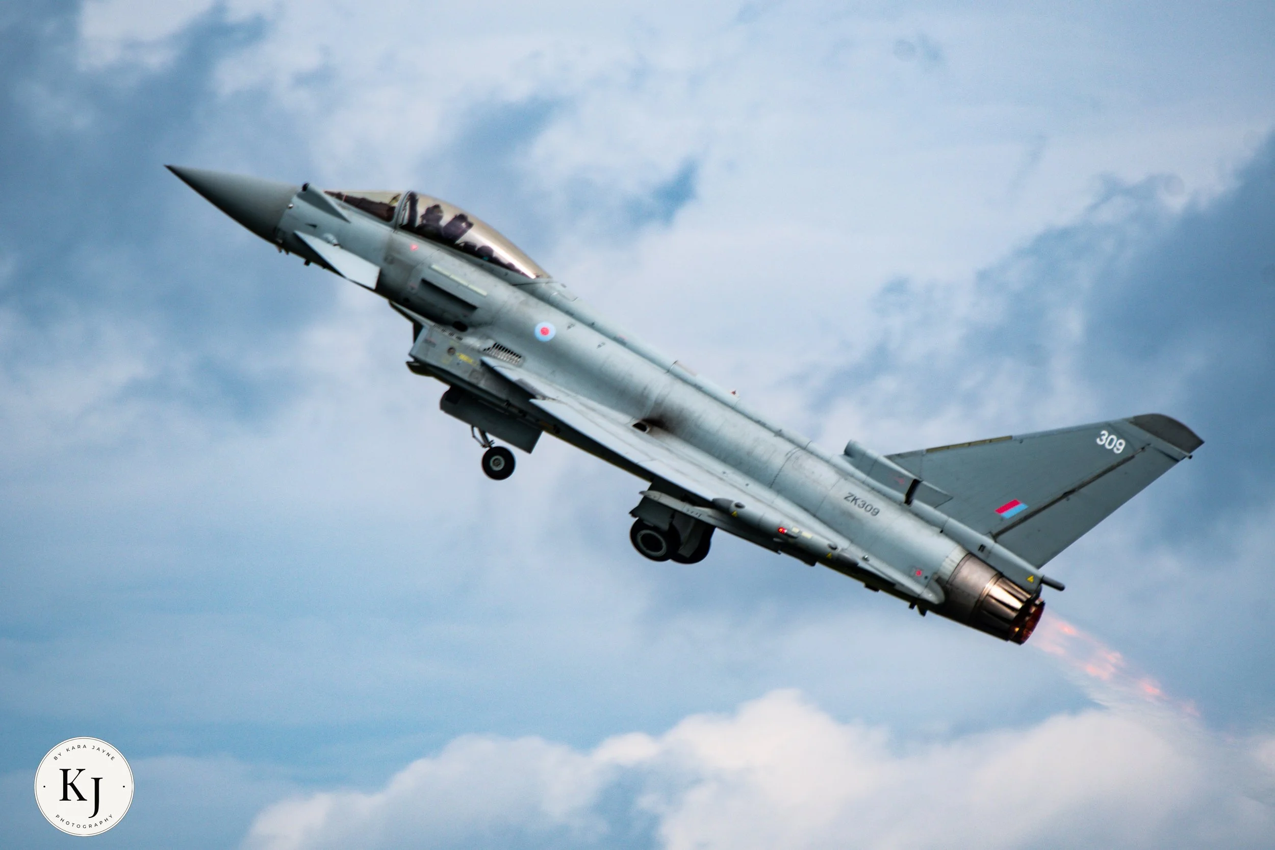 A military fighter jet flying in the sky with clouds visible in the background.