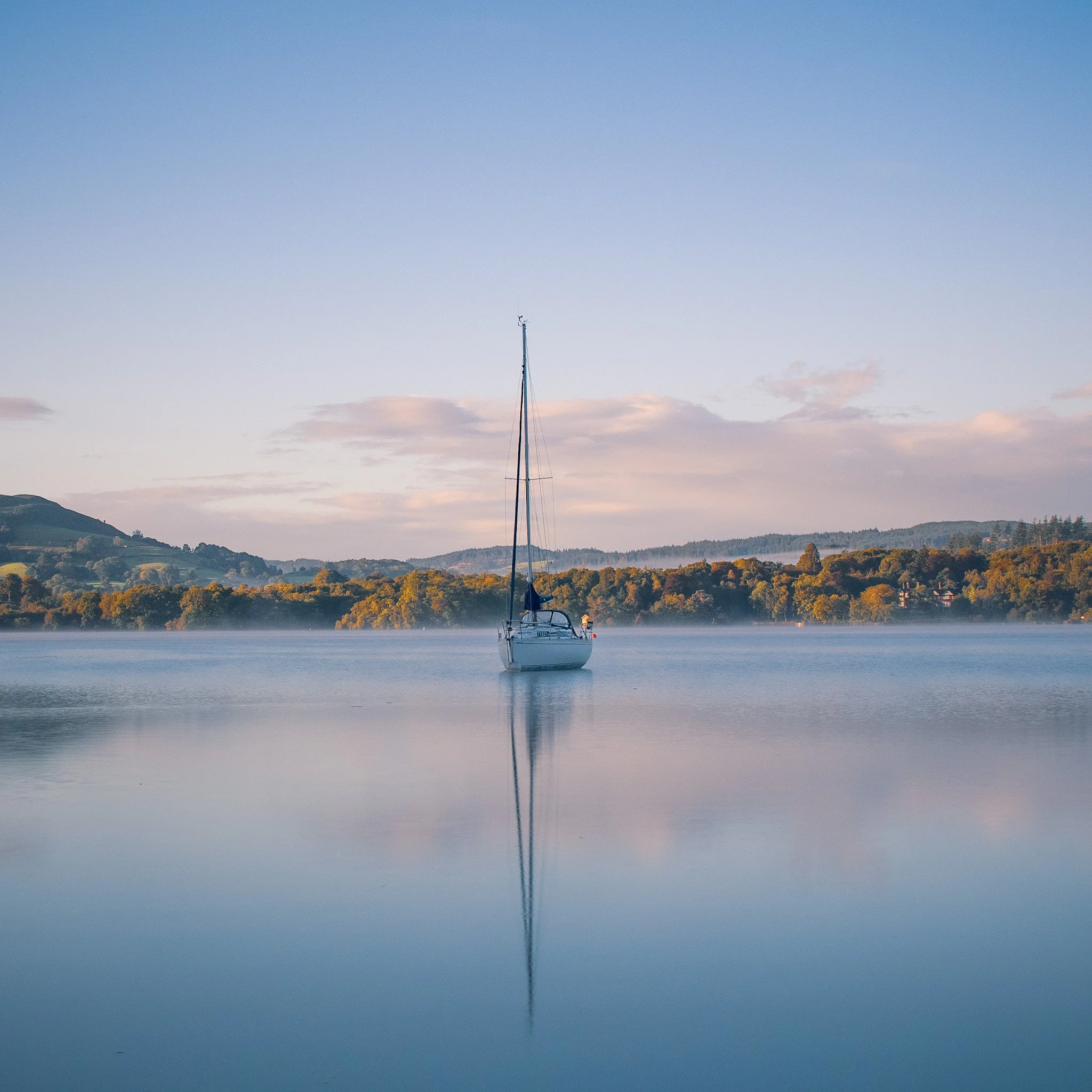 A sailboat floating on a calm body of water with a hilly landscape and trees in the background under a clear sky.