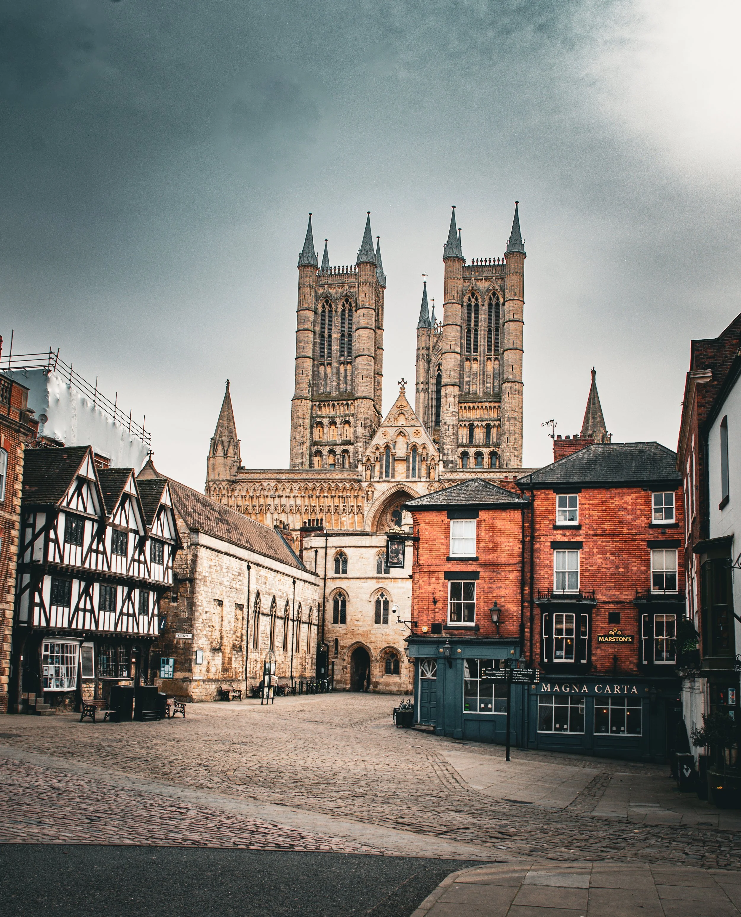 A historic city square with cobblestone pavement, surrounded by old buildings including a church with tall, pointed spires.