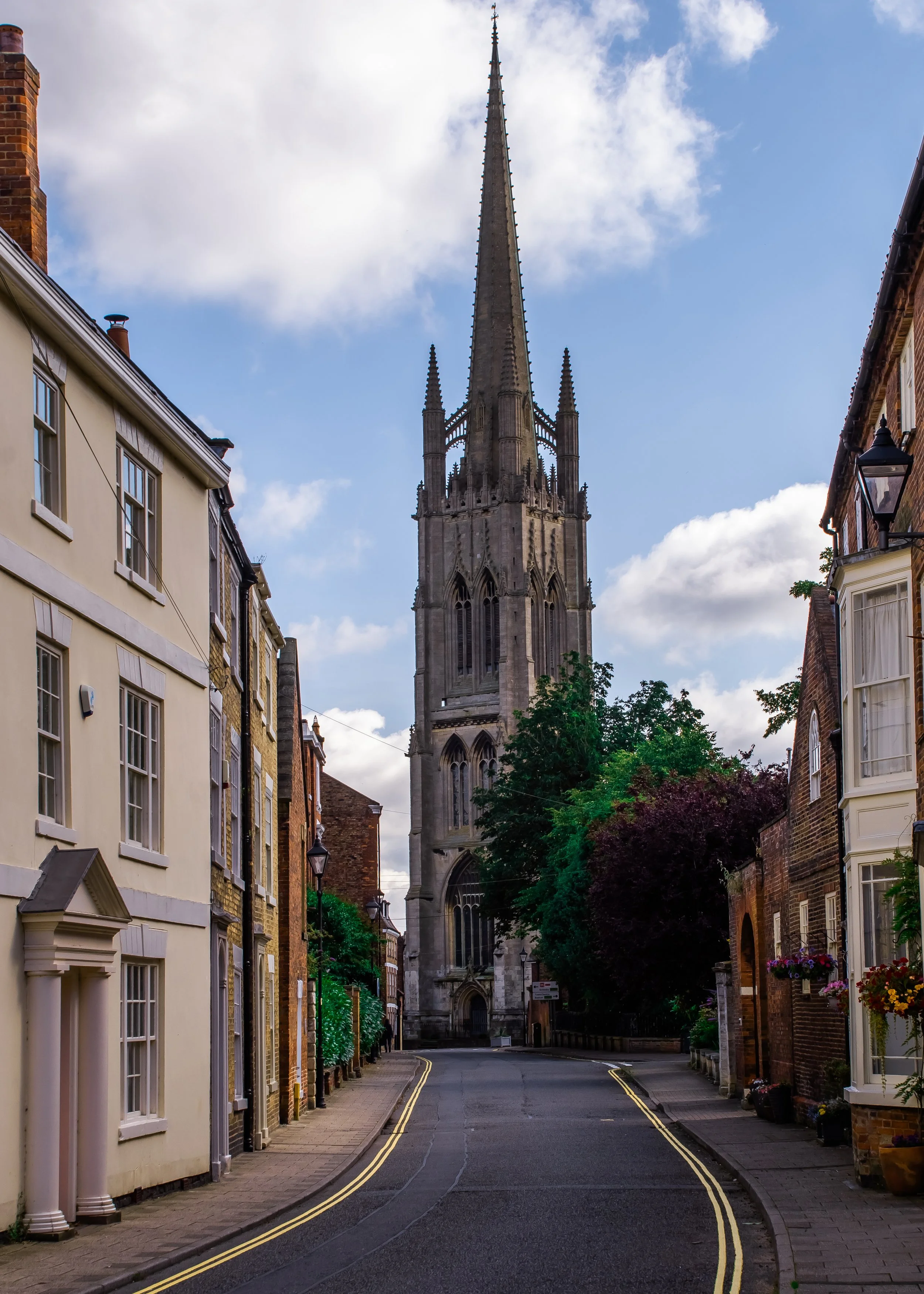 A deserted narrow street with buildings on both sides, and a tall church steeple in the distance, under a partly cloudy sky.