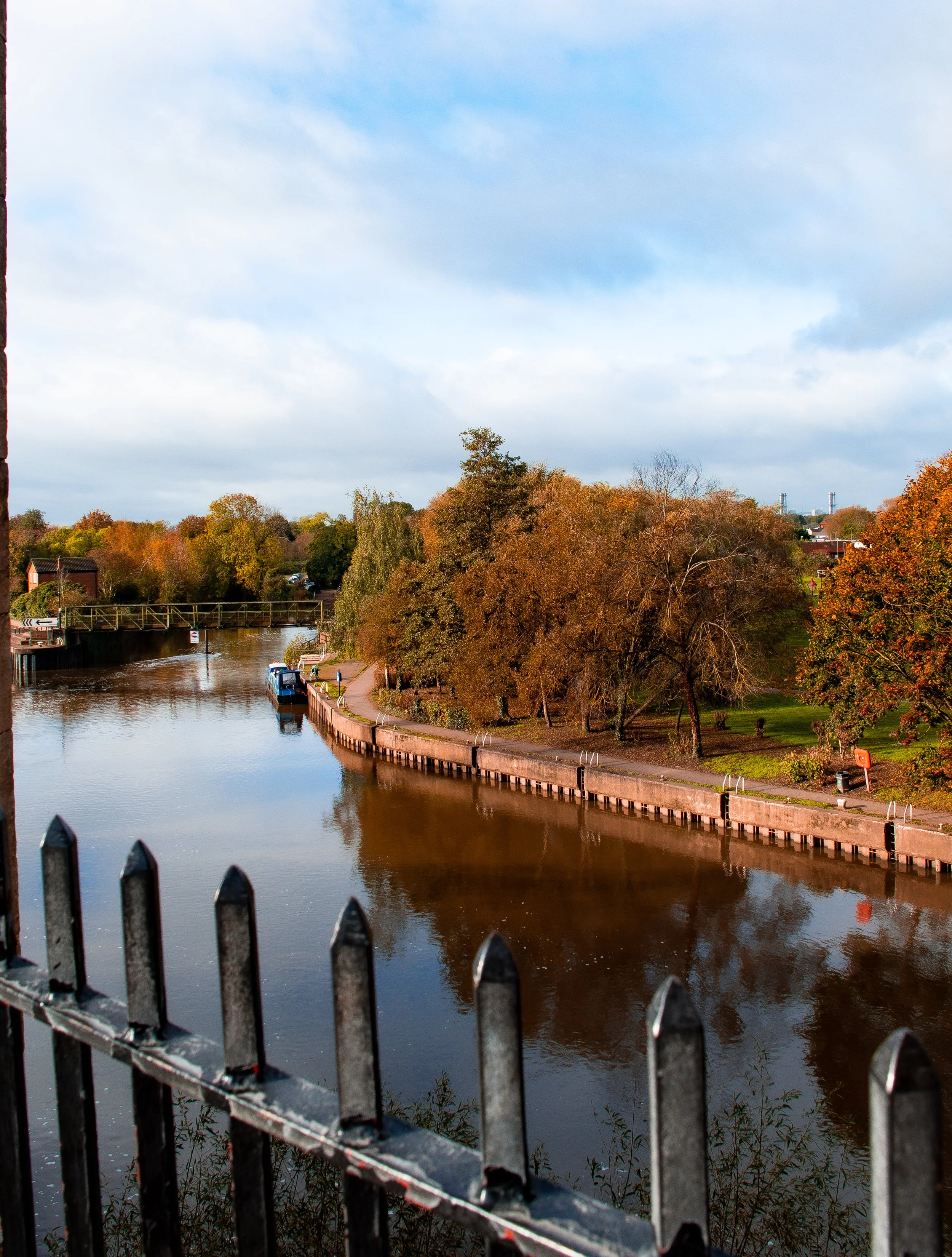Photo of a canal with apartment buildings, trees with autumn leaves, and a partly cloudy sky, taken through a metal fence.