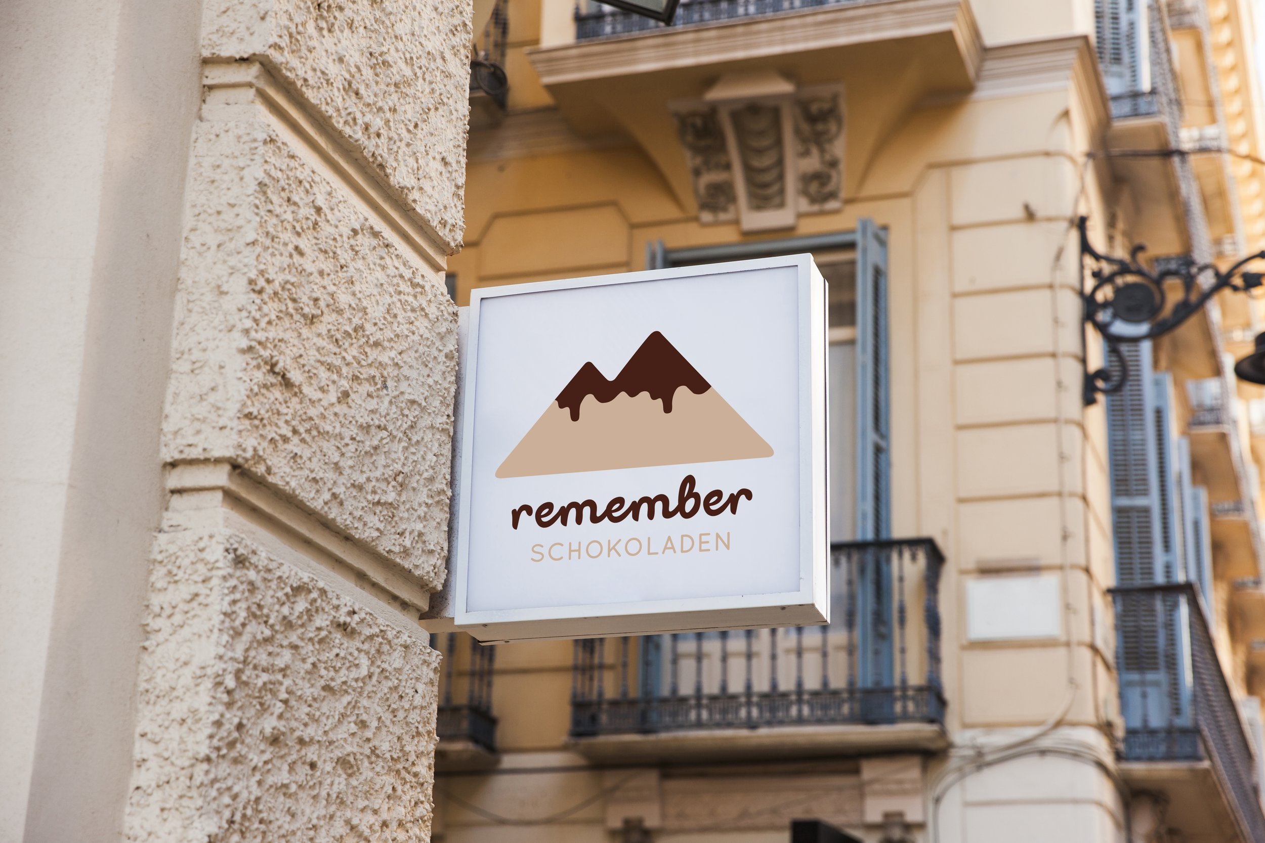 Straßenschild für einen Schokoladenladen namens 'Remember'. Das Schild ist an einer hellgelben, verzierten Gebäudefassade befestigt, mit mehreren Stockwerken und offenen Fenstern, einige mit schwarzen Eisenläden.