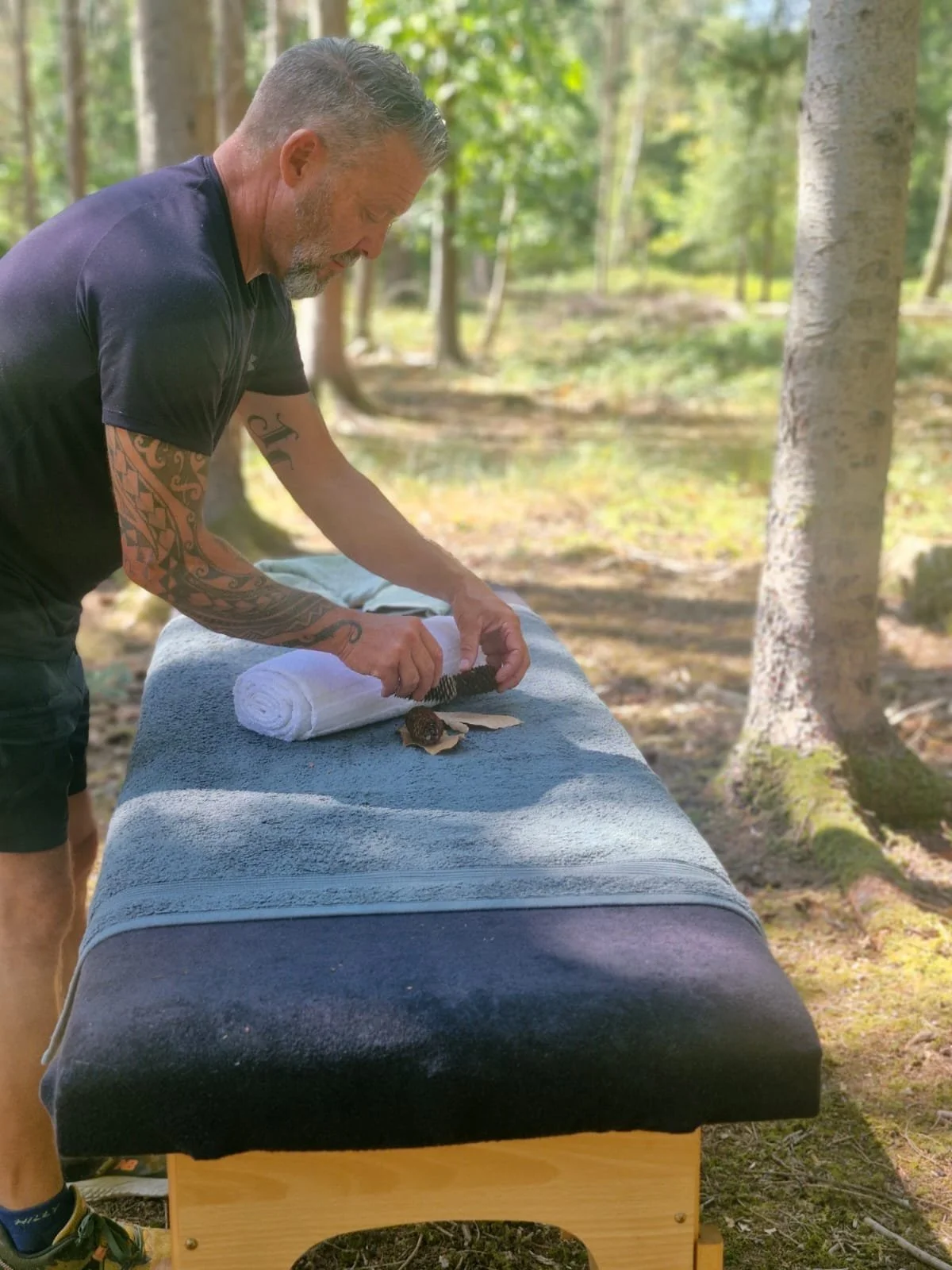 A man with tattoos on his arms is preparing a massage or spa treatment outdoors in a wooded area, using a brush on a wooden shell plant on a massage table.