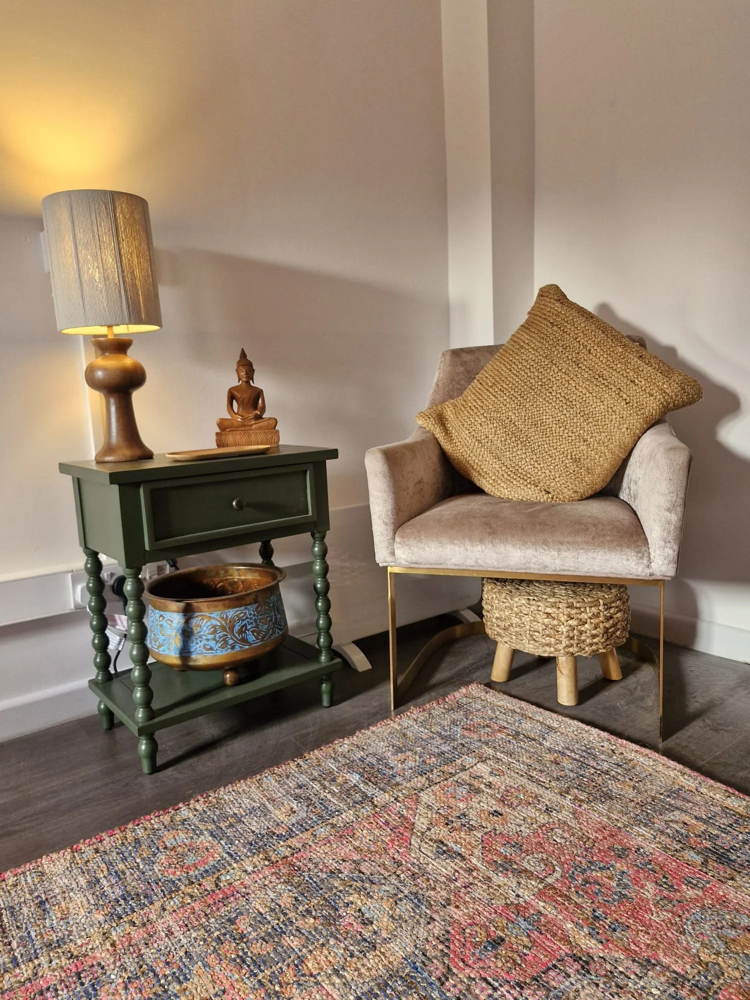 A cozy corner with a beige armchair, a gold pillow, a small green side table with a Buddha sculpture, a yellowish lamp, and a colorful patterned rug.