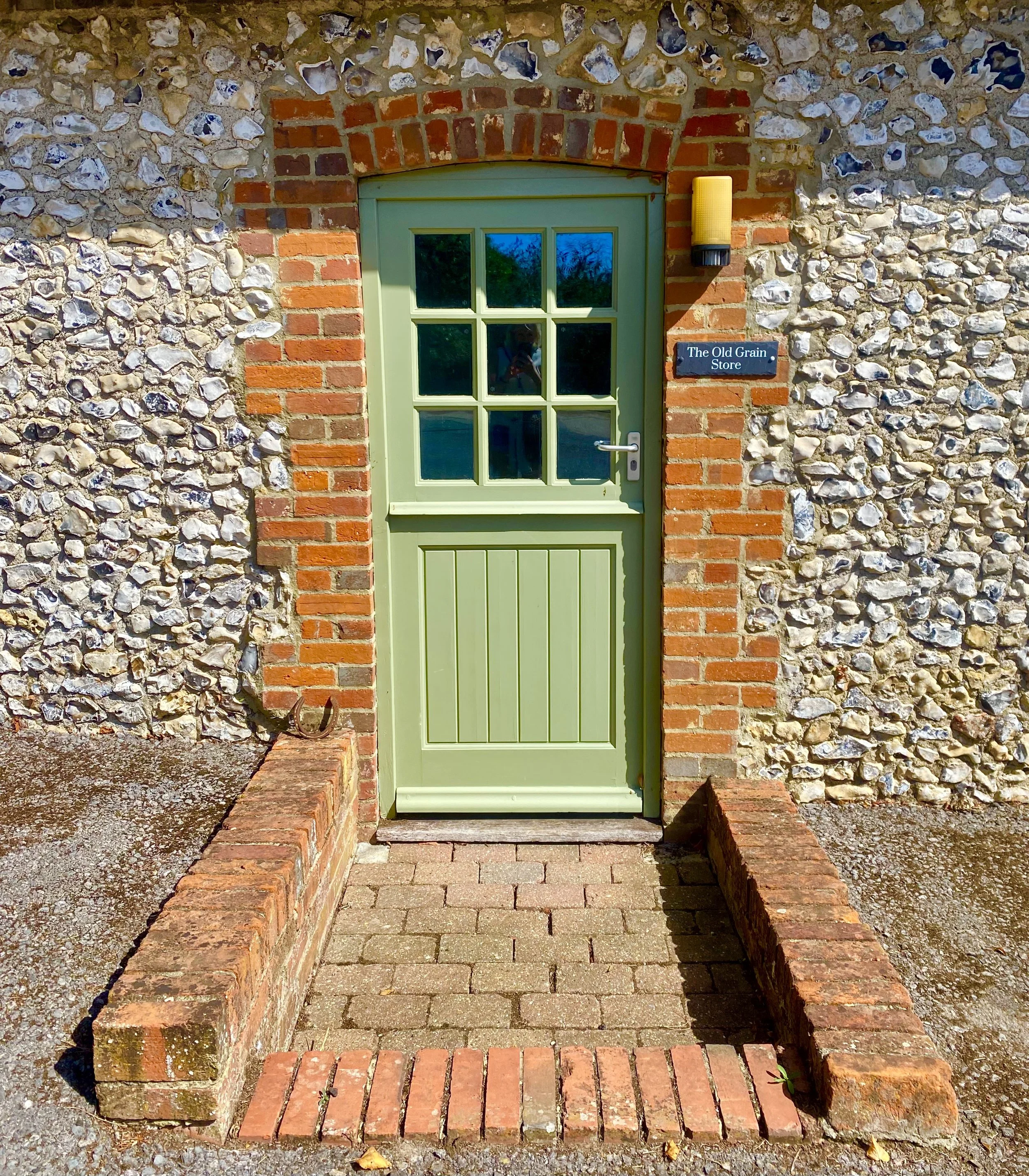 Green door with glass panes set in brick and stone wall, with a small brick step and a horse shoe placed on the step, and a black plaque to the right reading 'The Old Grain Store', under a yellow exterior light.
