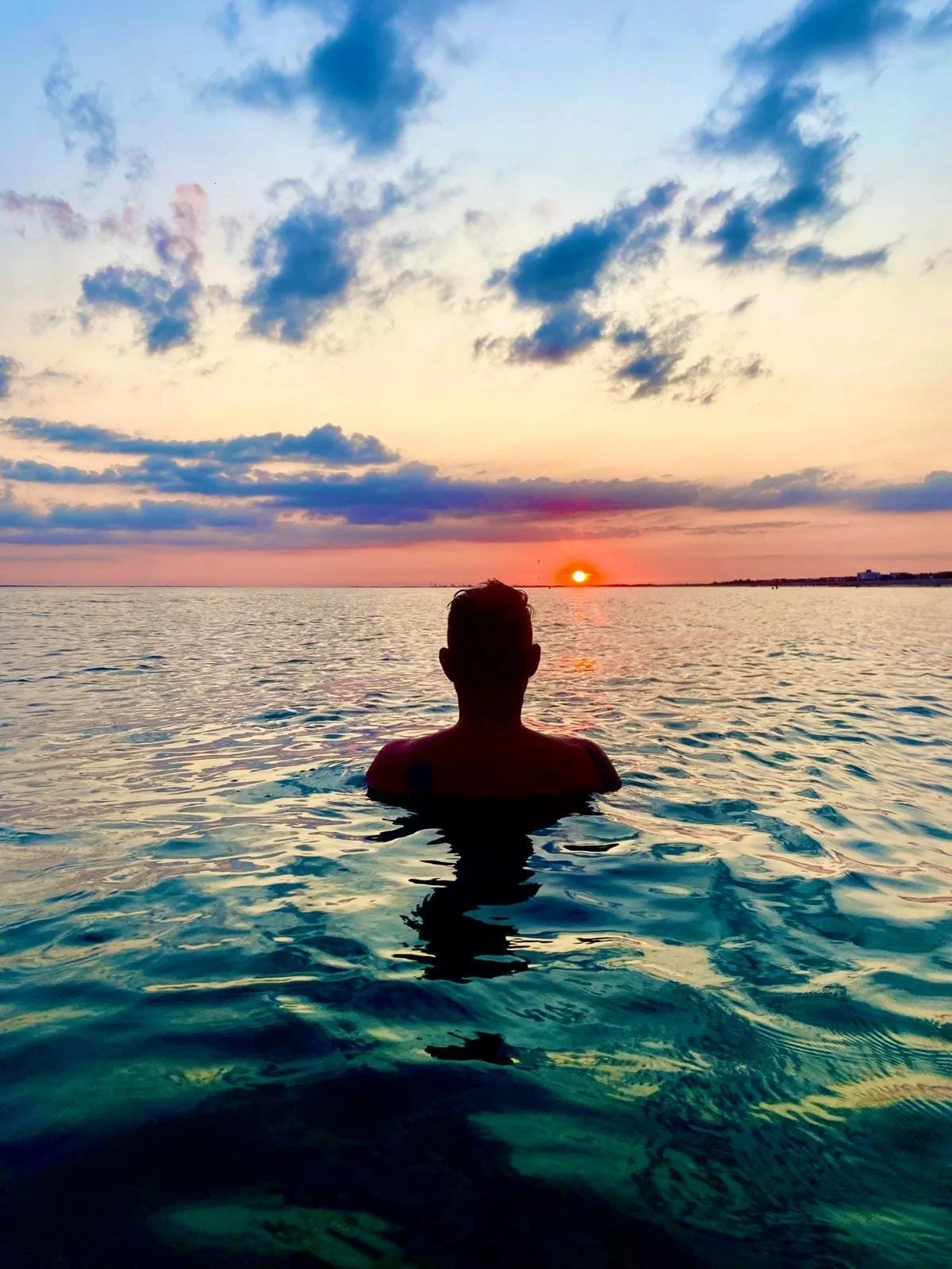 Person swimming in the ocean at sunset with colorful sky and clouds.