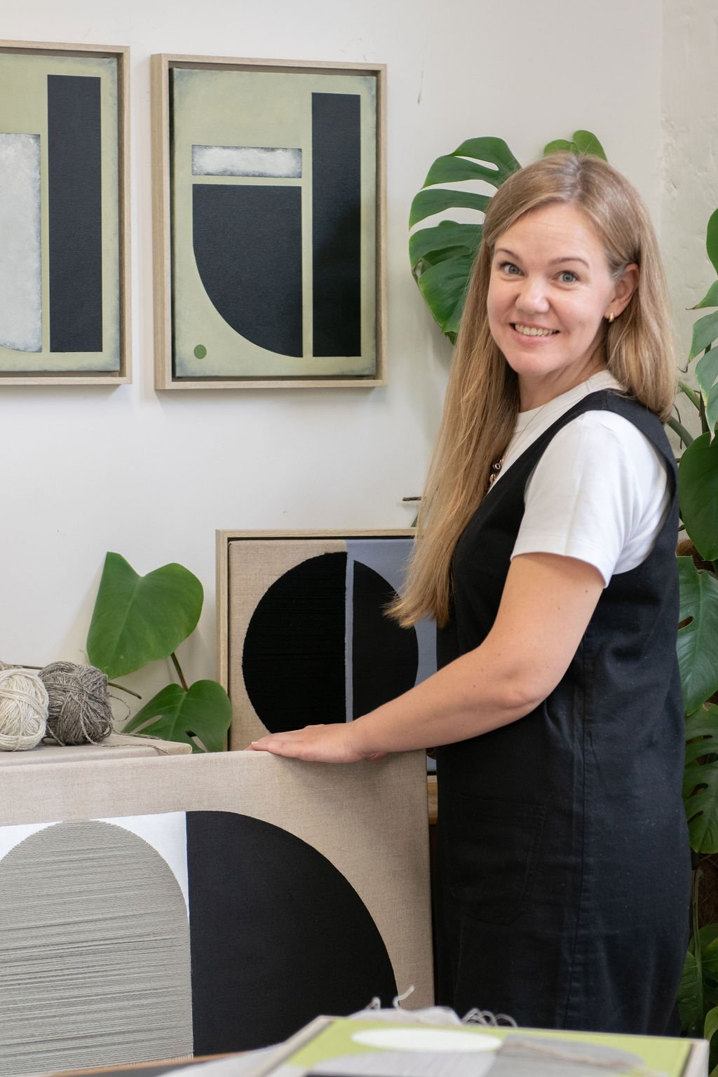 A woman, standing next to midcentury furniture piece with black and white geometric textile art next to it, in an art gallery or studio with framed geometric artwork on the wall and green plants nearby.