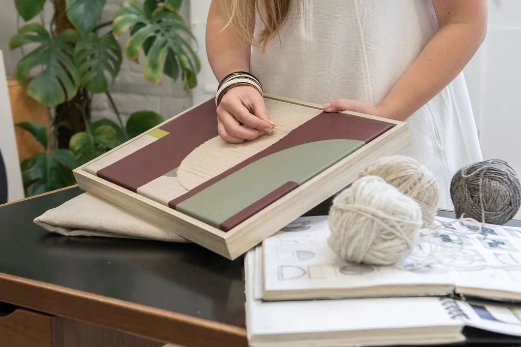 Person demonstrating a geometric art piece with yarn balls and a sketchbook on a table, greenery in the background.