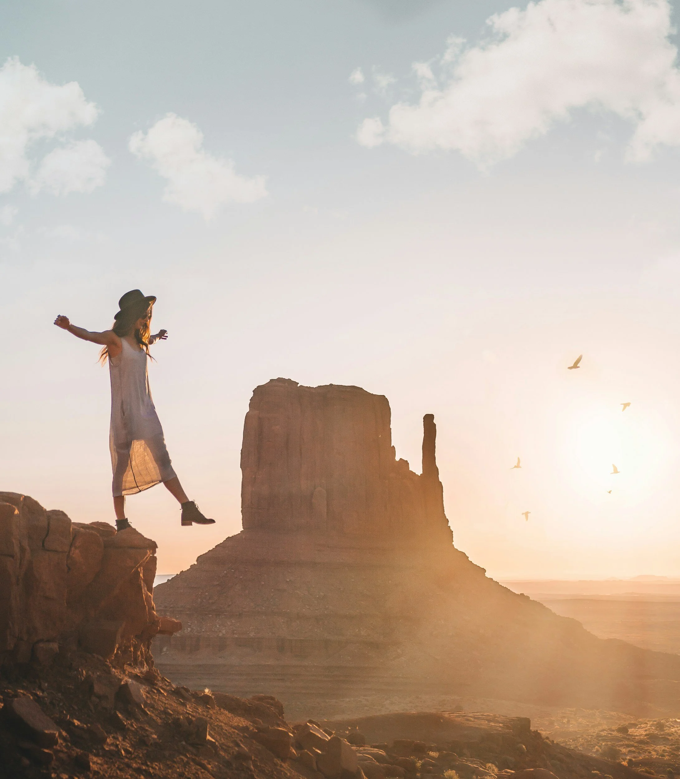 A woman with arms outstretched standing on rocky cliffs at sunset with ancient ruins and a flock of birds in the sky.