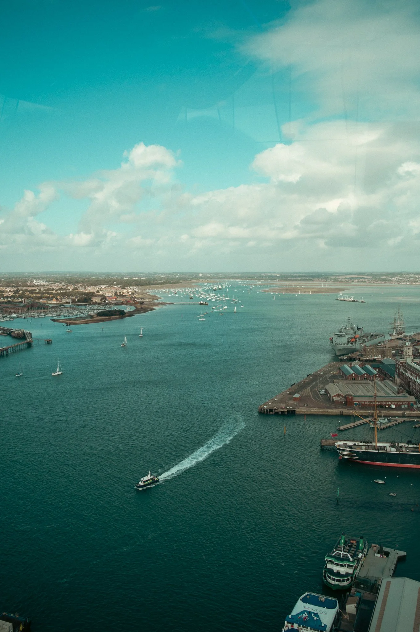 View of a harbor with sailboats, ships, and a motorboat leaving a wake, as seen from a high vantage point. Industrial and residential buildings line the shoreline, with a large body of water and a partly cloudy sky in the background.